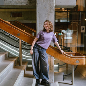 Model standing indoors on a staircase landing wearing Limited Edition Tees ’26 in mauve featuring a small white RMIT University logo printed on the left chest, paired with loose dark denim wide-leg jeans and black lace-up boots, leaning against a wooden handrail with industrial concrete and escalator background, available at the RMIT Store.