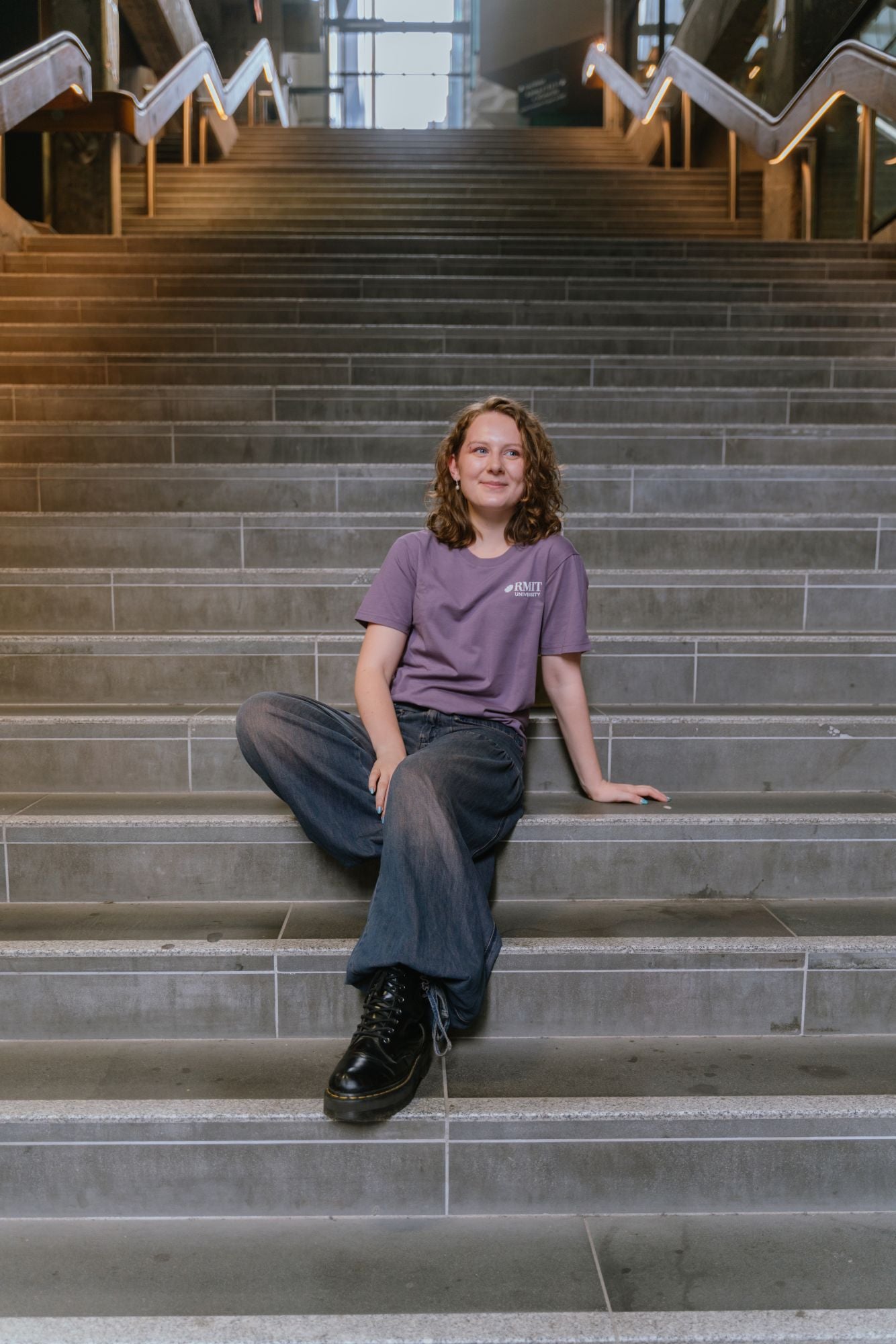 Model seated on wide indoor concrete stairs wearing Limited Edition Tees ’26 in mauve with a small white RMIT University logo on the left chest, styled with dark denim wide-leg jeans and black boots, sitting relaxed with one arm resting on the step in a modern stairwell setting, available at the RMIT Store.