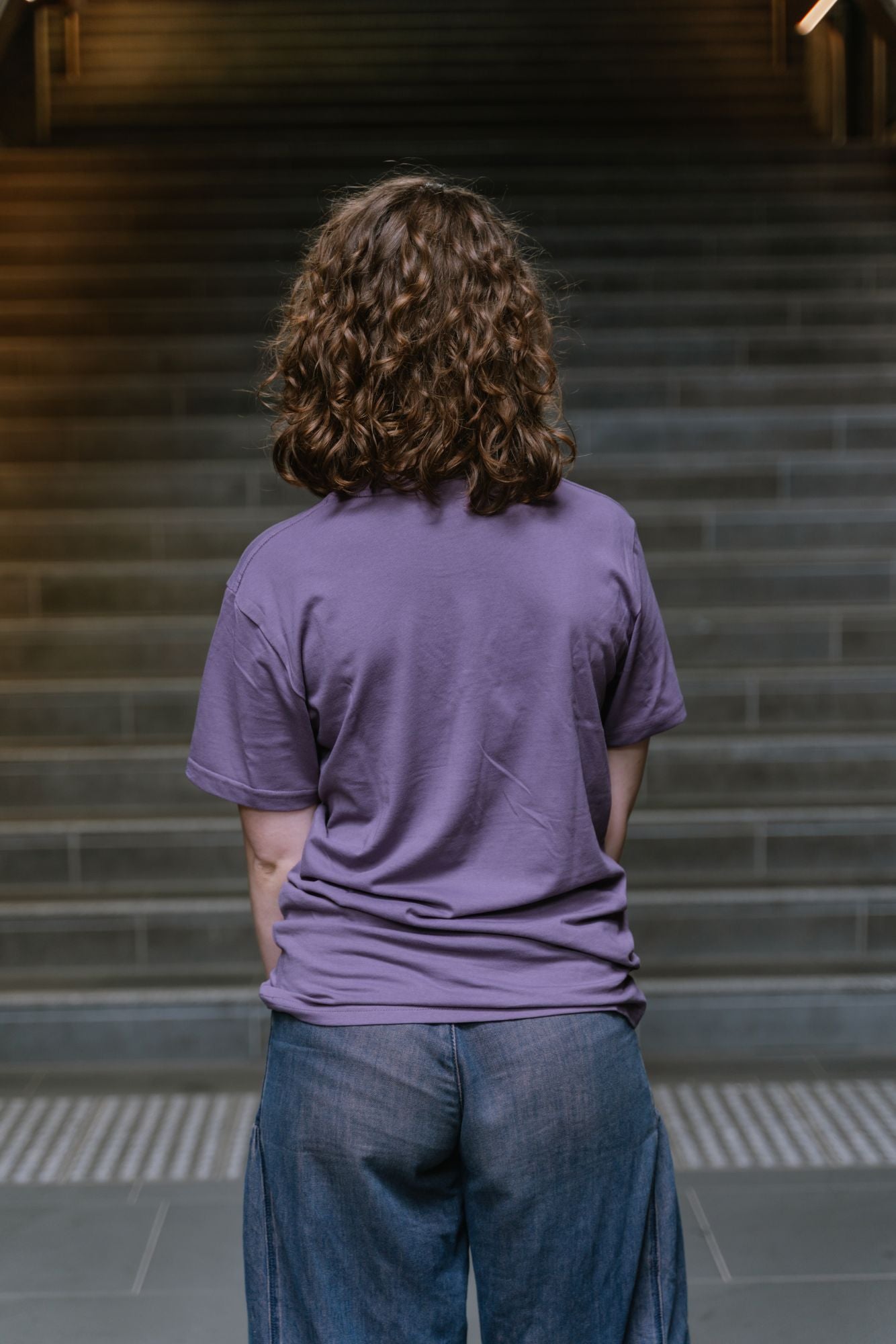 Rear view of model standing indoors on staircase wearing Limited Edition Tees ’26 in mauve showing plain back with no visible print, paired with dark denim wide-leg jeans, with soft lighting and blurred stair background, available at the RMIT Store.