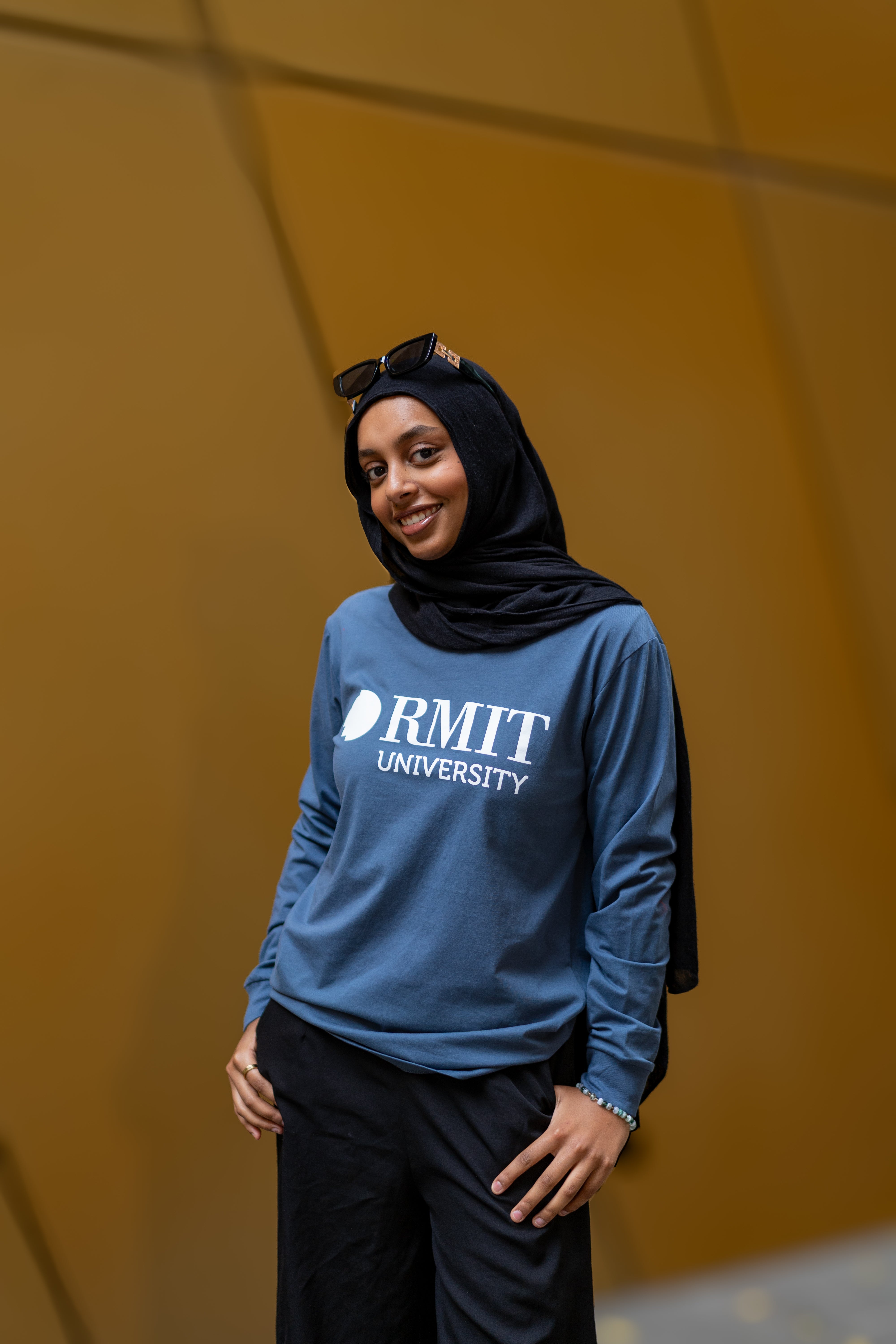 Model wearing RMIT Loud Logo Long Sleeve Tee in slate blue, paired with flowing black trousers and accessorised with a black headscarf and sunglasses on the head.