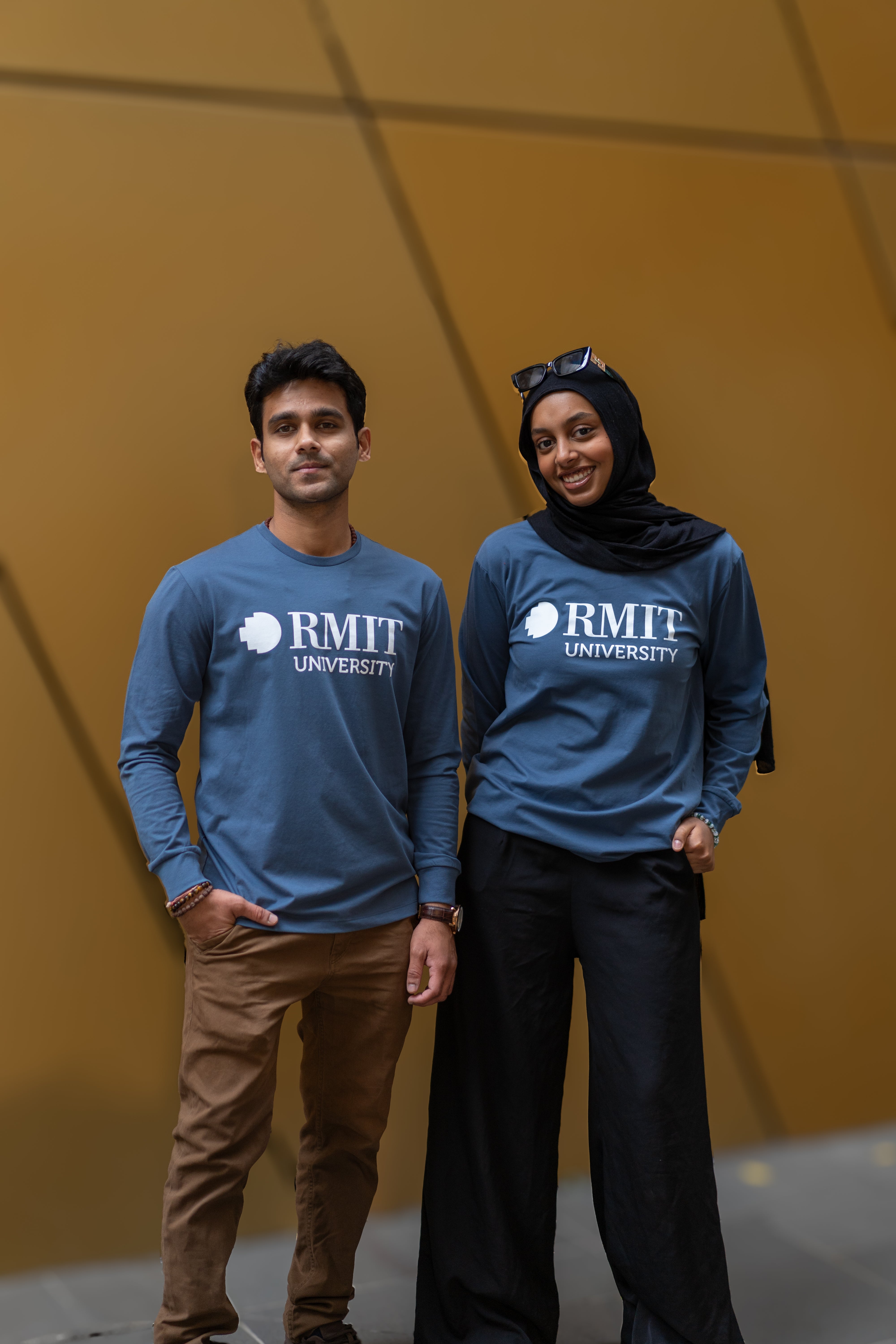 Two models showcasing RMIT Loud Logo Long Sleeve Tees in slate blue; the left model in light brown trousers, the right model in black, both with casual wrist accessories.
