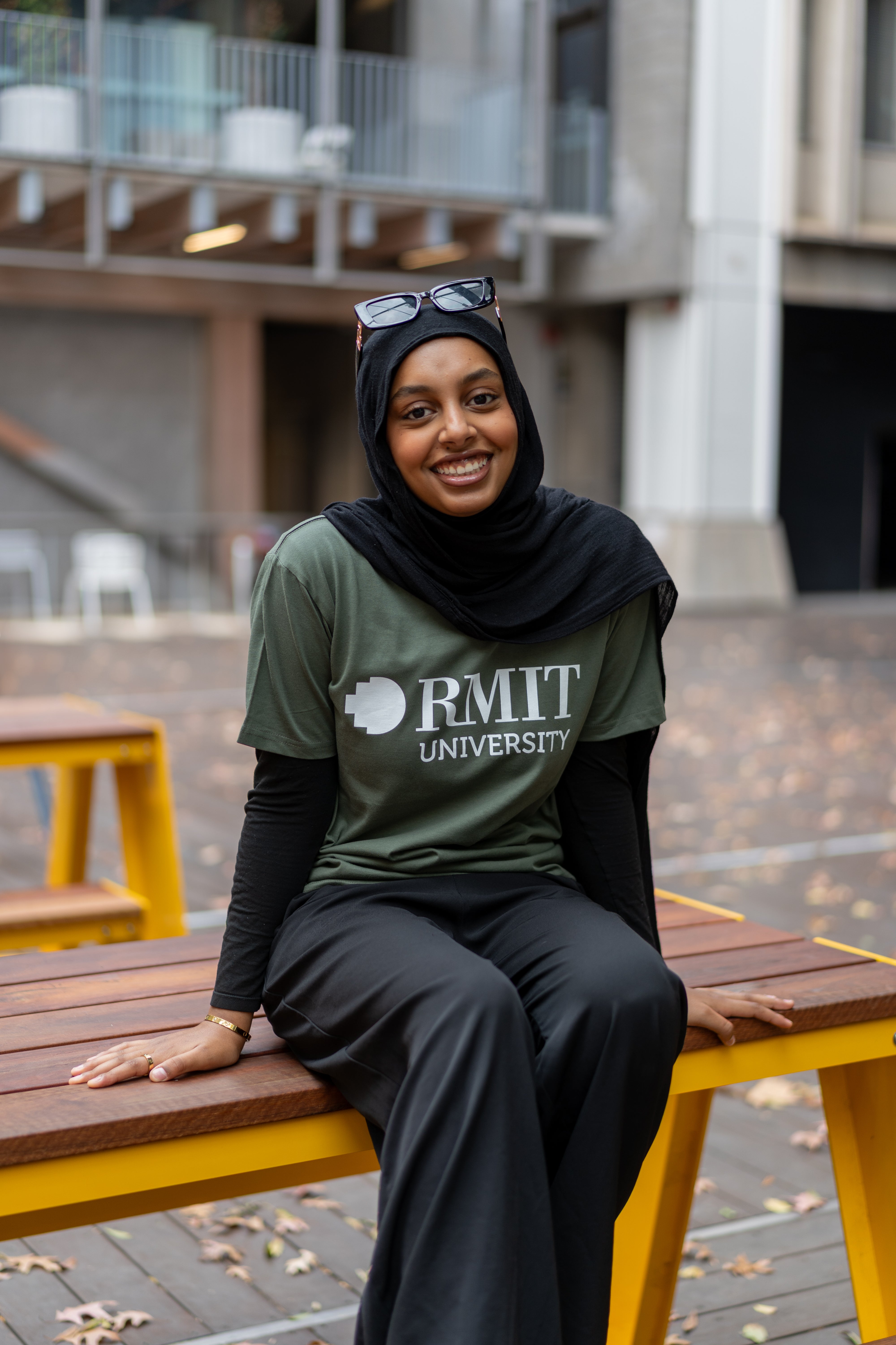 Model comfortably sitting on a bench, showcasing the RMIT Store Loud Logo Short Sleeve Tee in cypress. The ensemble includes black ankle-length trousers and subtle wrist accessories.
