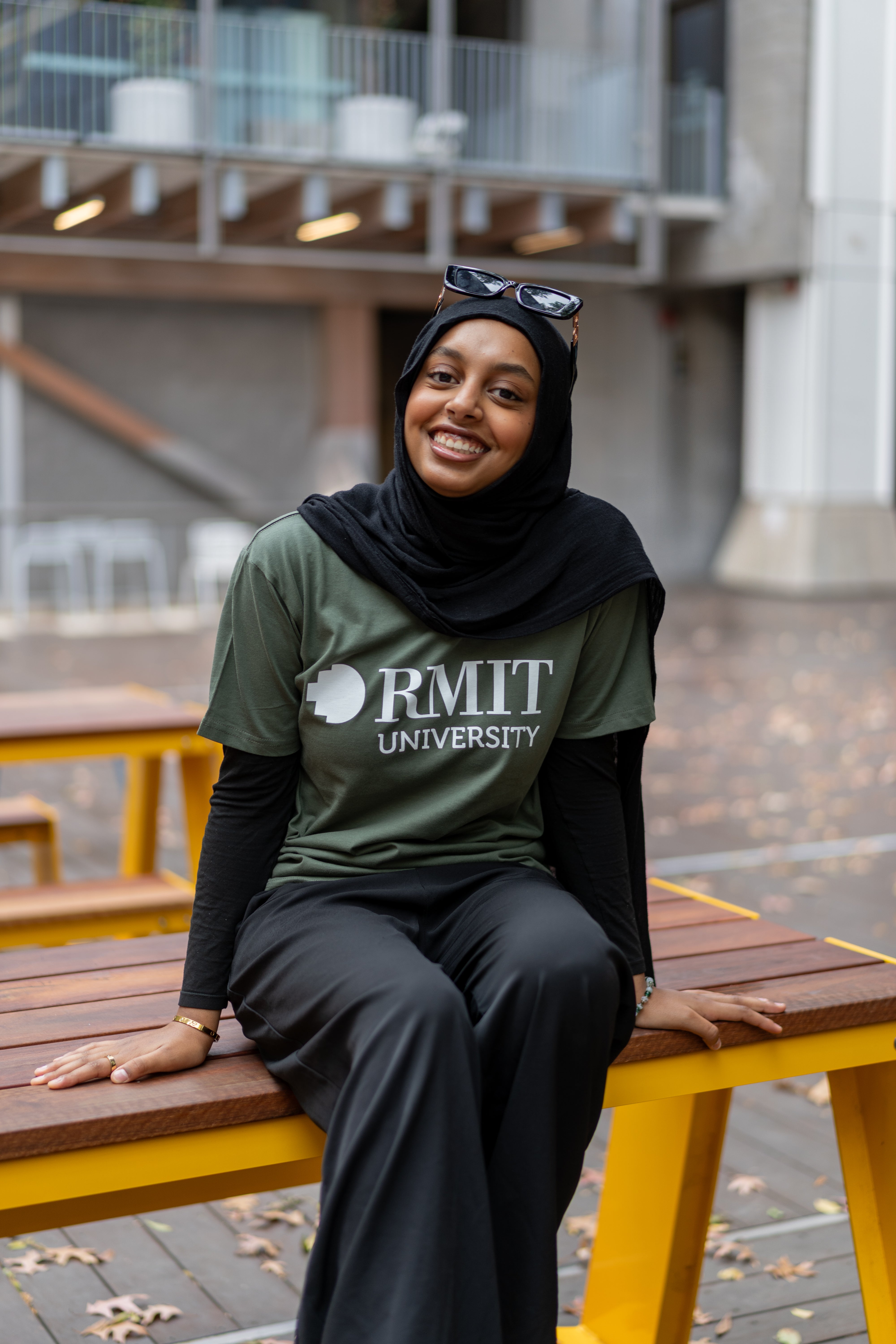 Model seated on a wooden bench wearing the RMIT Store Loud Logo Short Sleeve Tee in cypress, paired with black trousers and a black headscarf. The tee features a prominent white RMIT University logo.