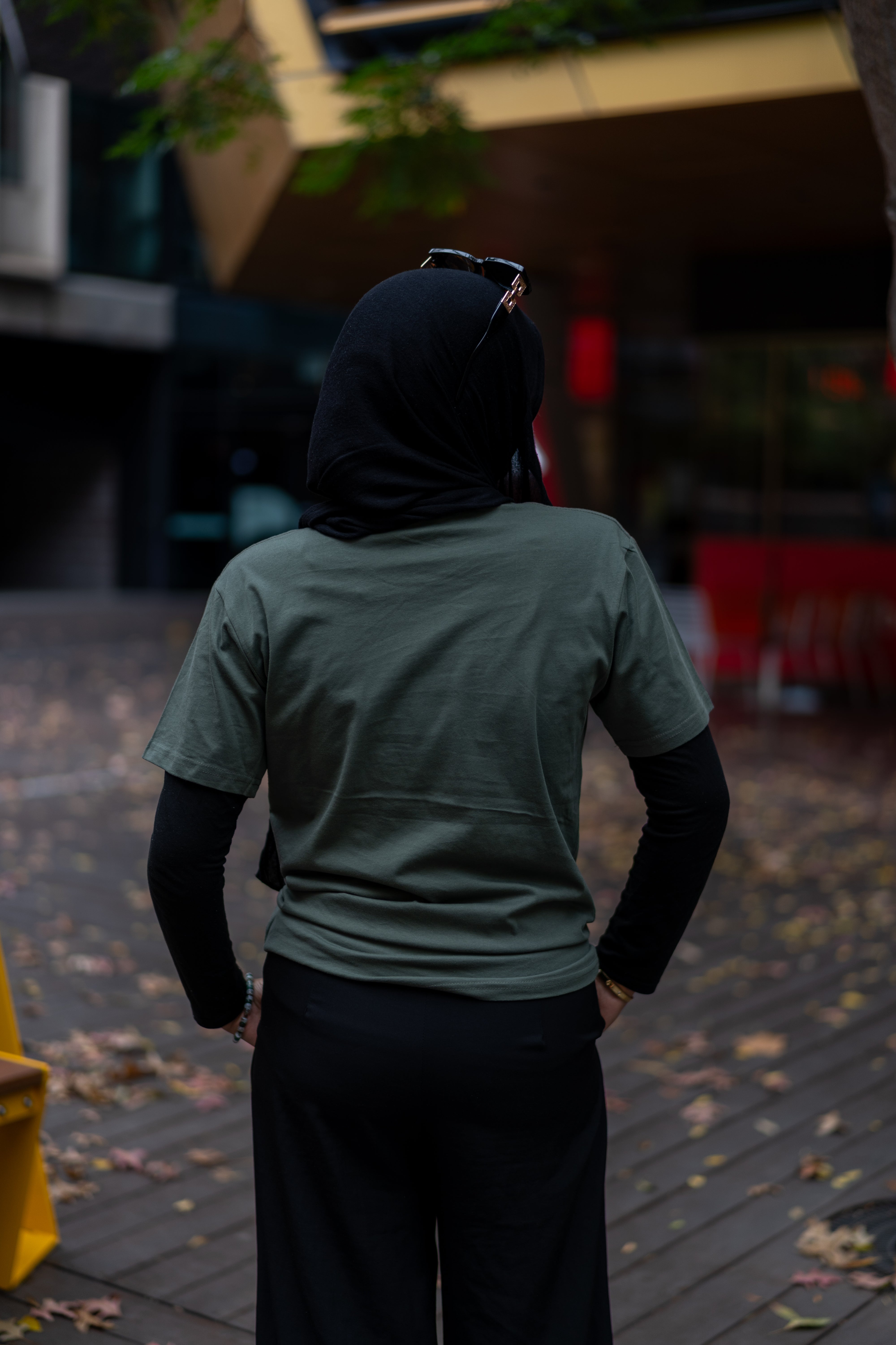 Rear view of a model wearing the RMIT Store Loud Logo Short Sleeve Tee in cypress, styled with a long-sleeve black undershirt and black headscarf, in an urban outdoor setting.