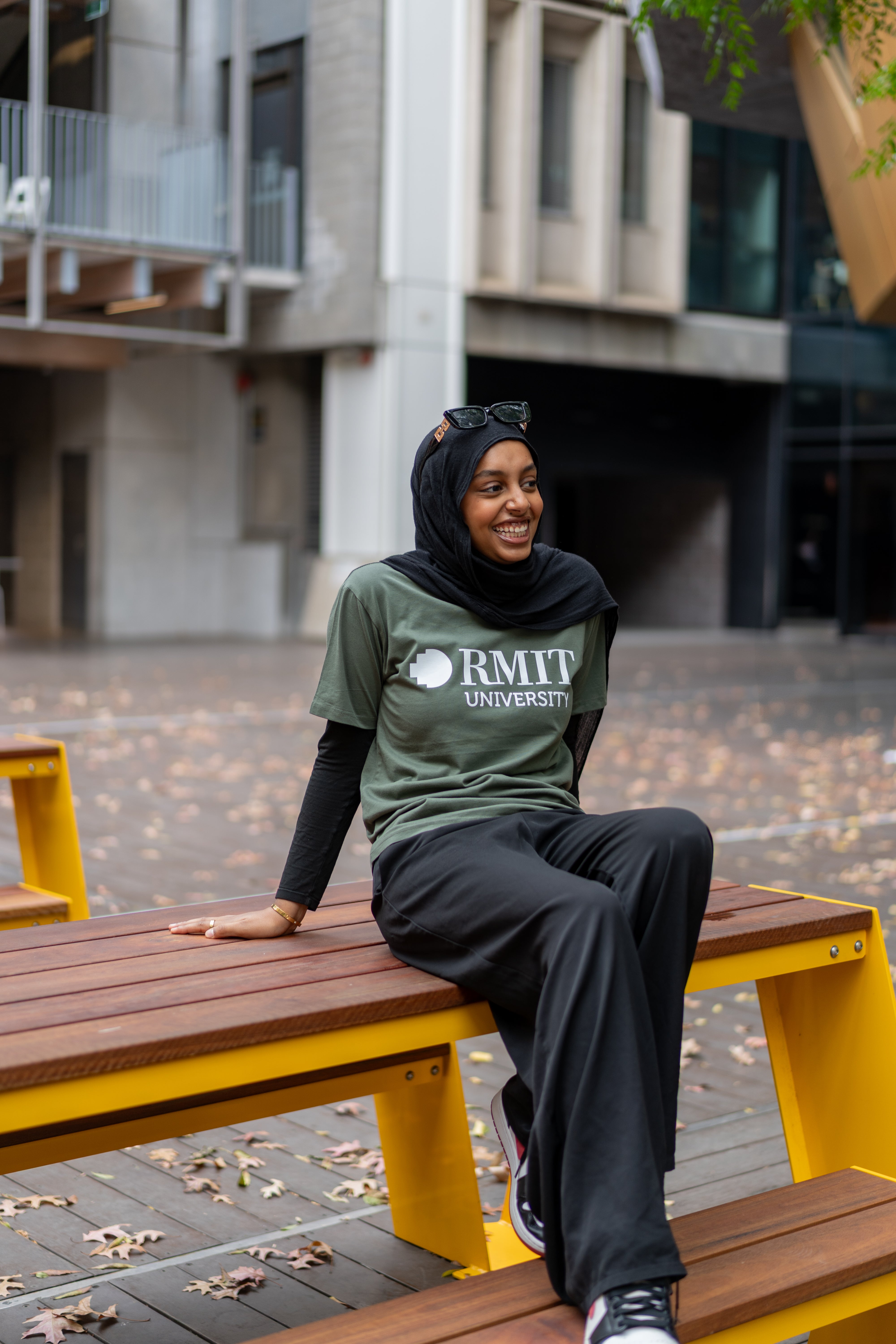 Model poses on a bright yellow wooden bench, dressed in RMIT Store Loud Logo Short Sleeve Tee in cypress. The look is completed with black trousers, black sneakers, and sunglasses resting on the head.