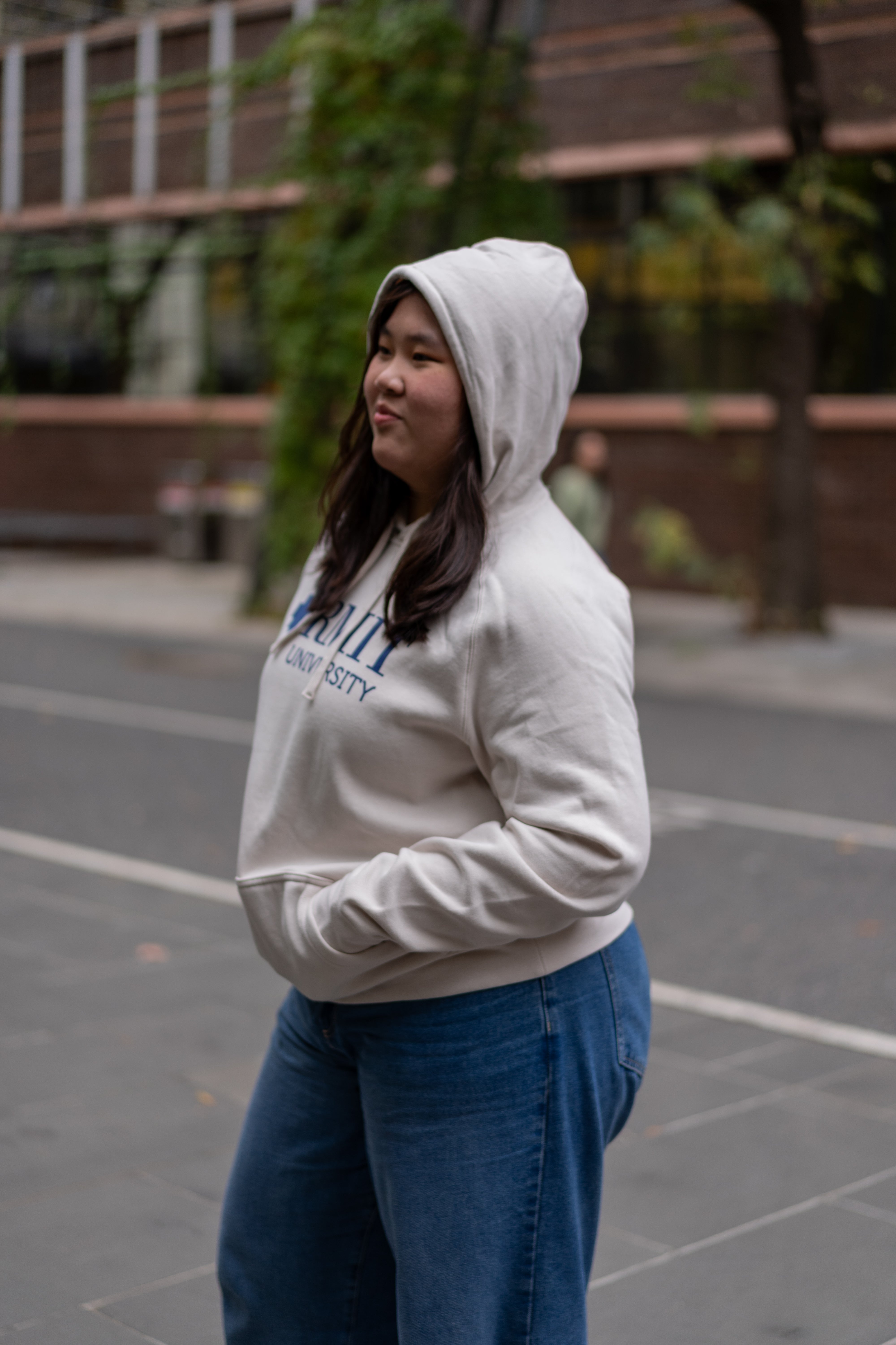 A model wears the RMIT Ltd Ed Hoodie in bone, featuring a side view. The hoodie displays an embroidered "RMIT University" logo in blue. Crafted from soft and high-quality material, this hoodie combines comfort with style, available at the RMIT Store.