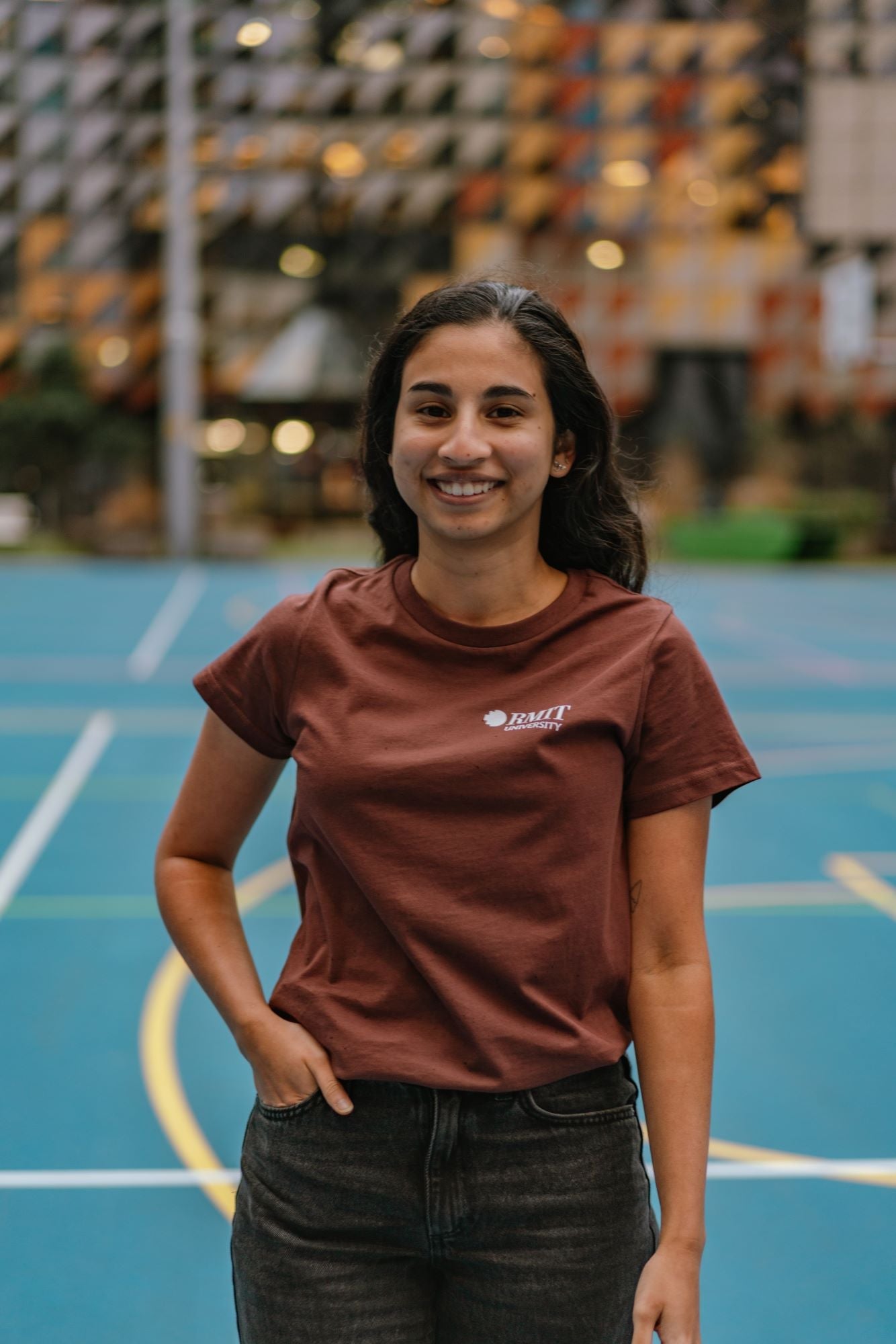 RMIT Limited Edition Tee in chestnut brown, featuring a small white RMIT University logo on the left chest. Female model standing in a campus setting, smiling softly with one arm relaxed at her side and the other arm in her pocket. Available at the RMIT Store.