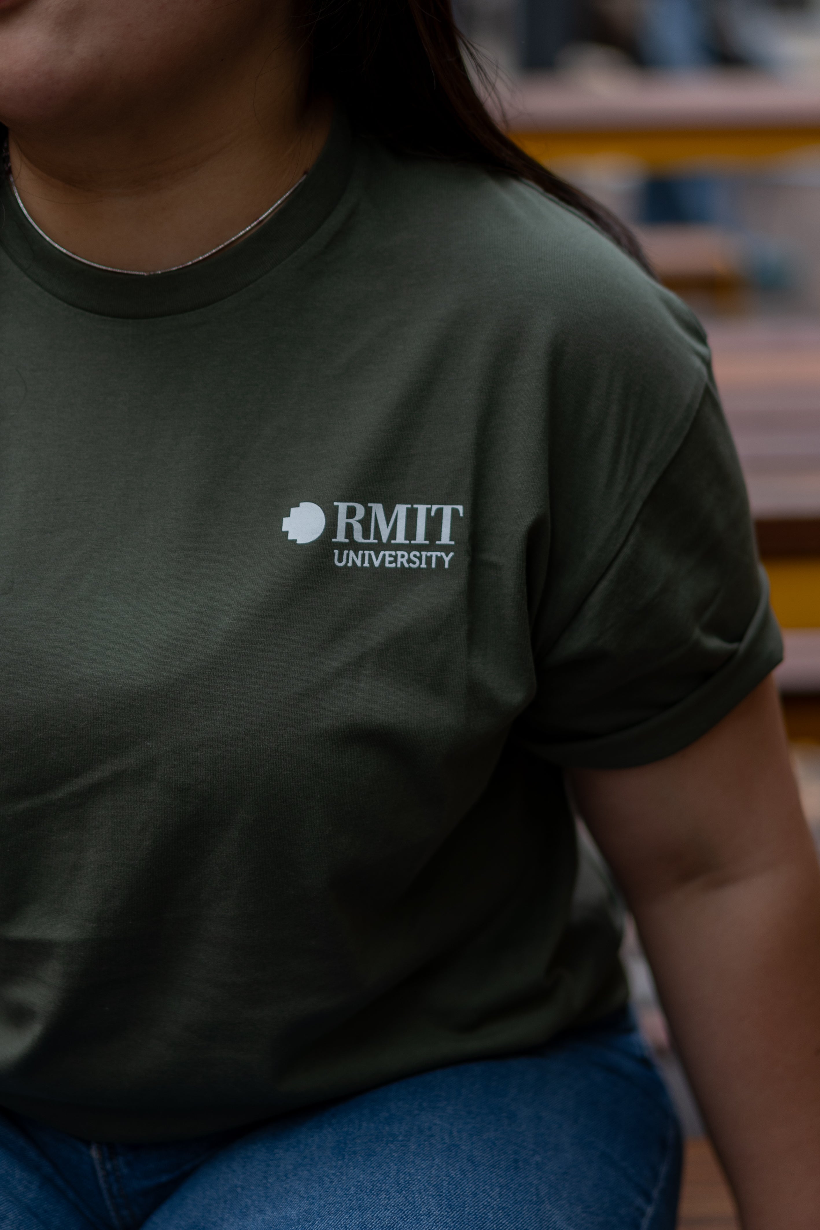 Close-up of model wearing the RMIT Ltd Ed Tee in Cypress green, featuring the RMIT University logo printed on the chest. Styled with casual blue jeans, available at the RMIT Store.