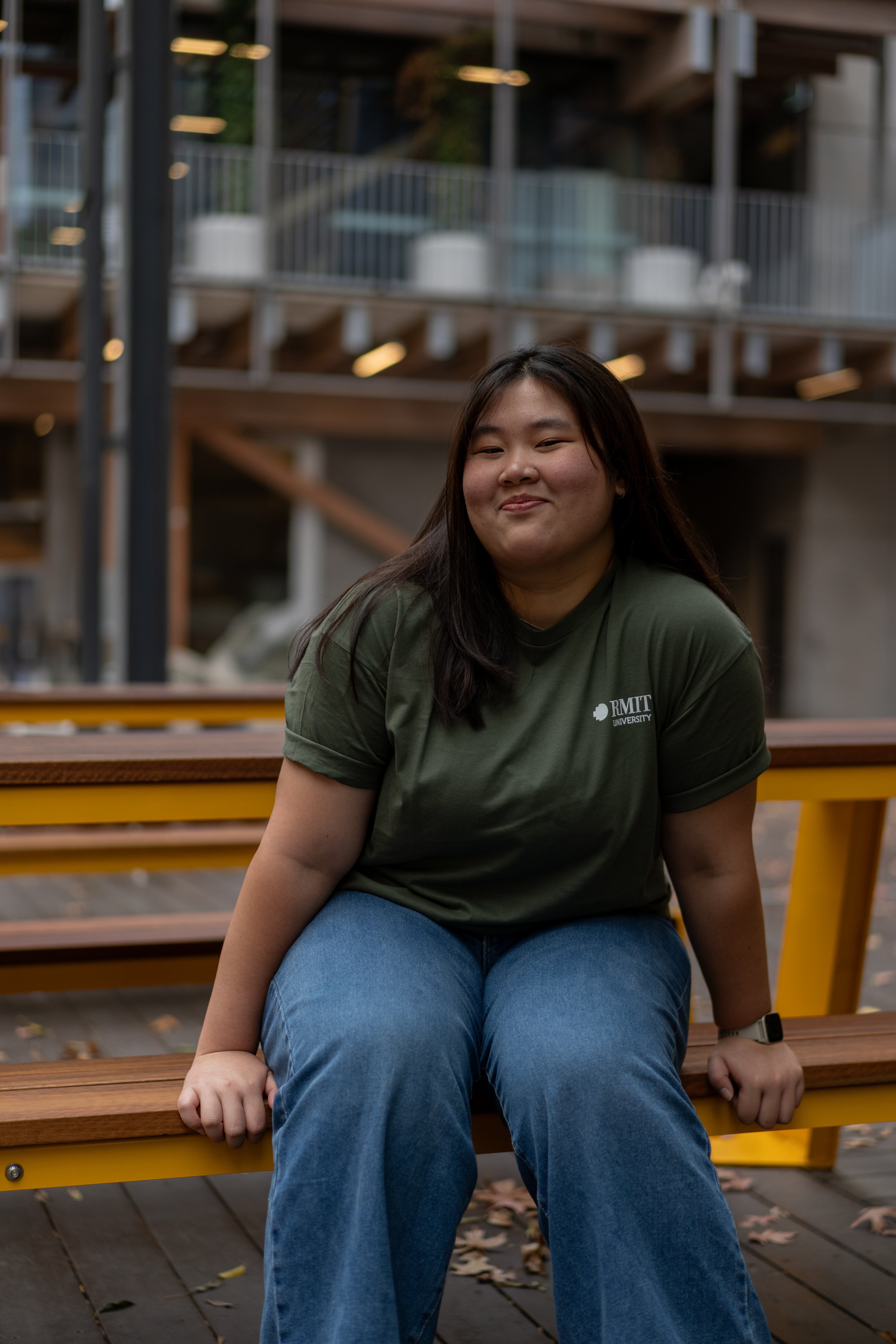 Model wears the RMIT Ltd Ed Tee in Cypress green, seated on wooden outdoor benches. Paired with blue jeans for a relaxed look, available at the RMIT Store.