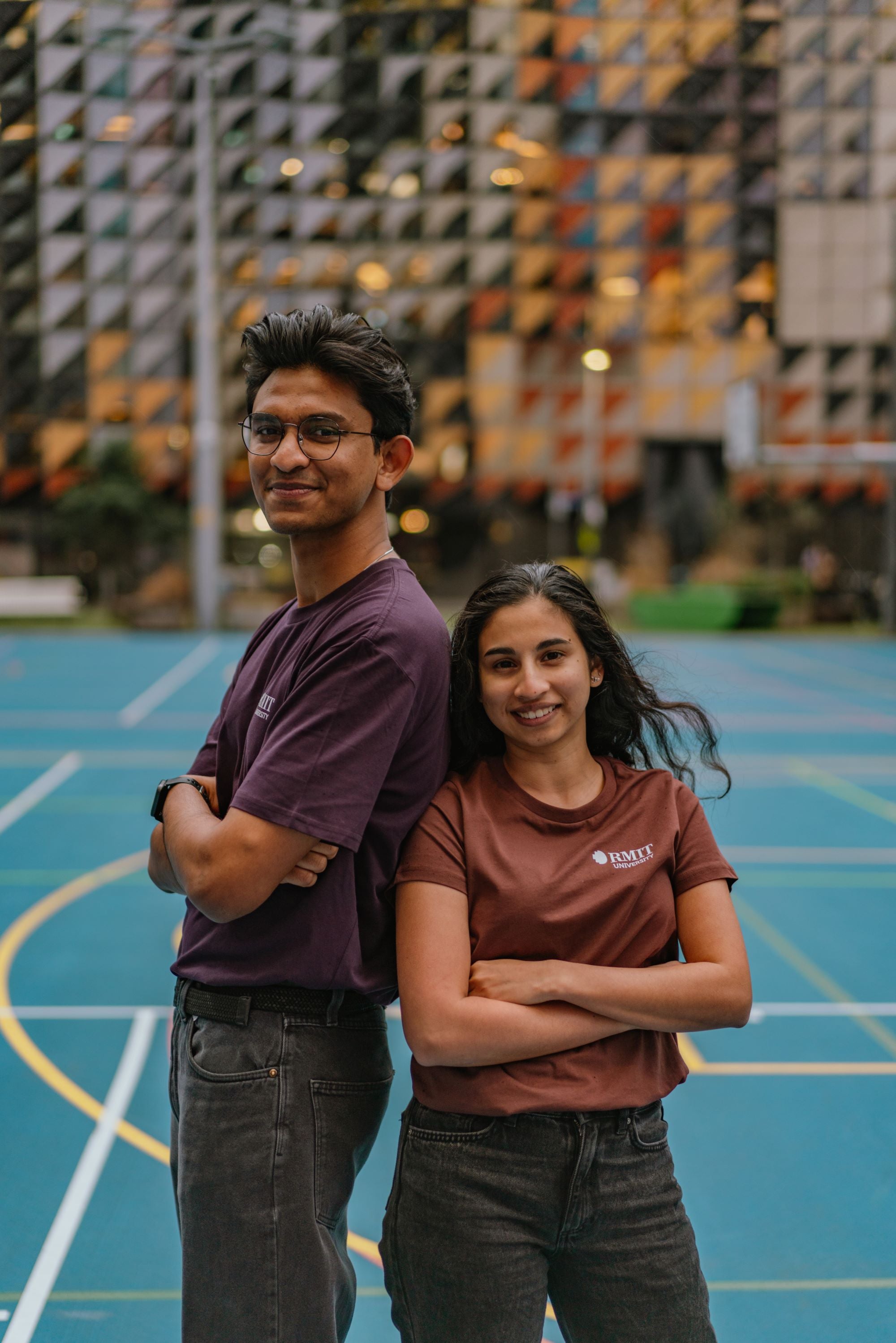 RMIT Limited Edition Tees in purple and chestnut worn by a male and female model standing back-to-back outdoors in a campus setting. The male model wears the purple variant and the female wears the chestnut; both are looking into the camera with relaxed expressions. Each tee features a small white RMIT University logo on the left chest. Available at the RMIT Store.