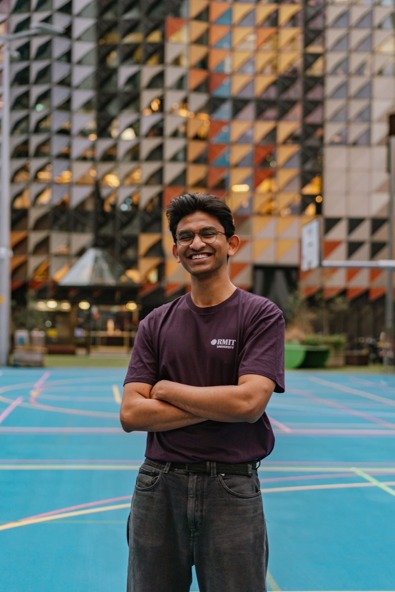 RMIT Limited Edition Tee in purple, featuring a small white RMIT University logo on the left chest. A male model is wearing it and standing in a campus setting with arms crossed over his chest and smiling. Available at the RMIT Store.