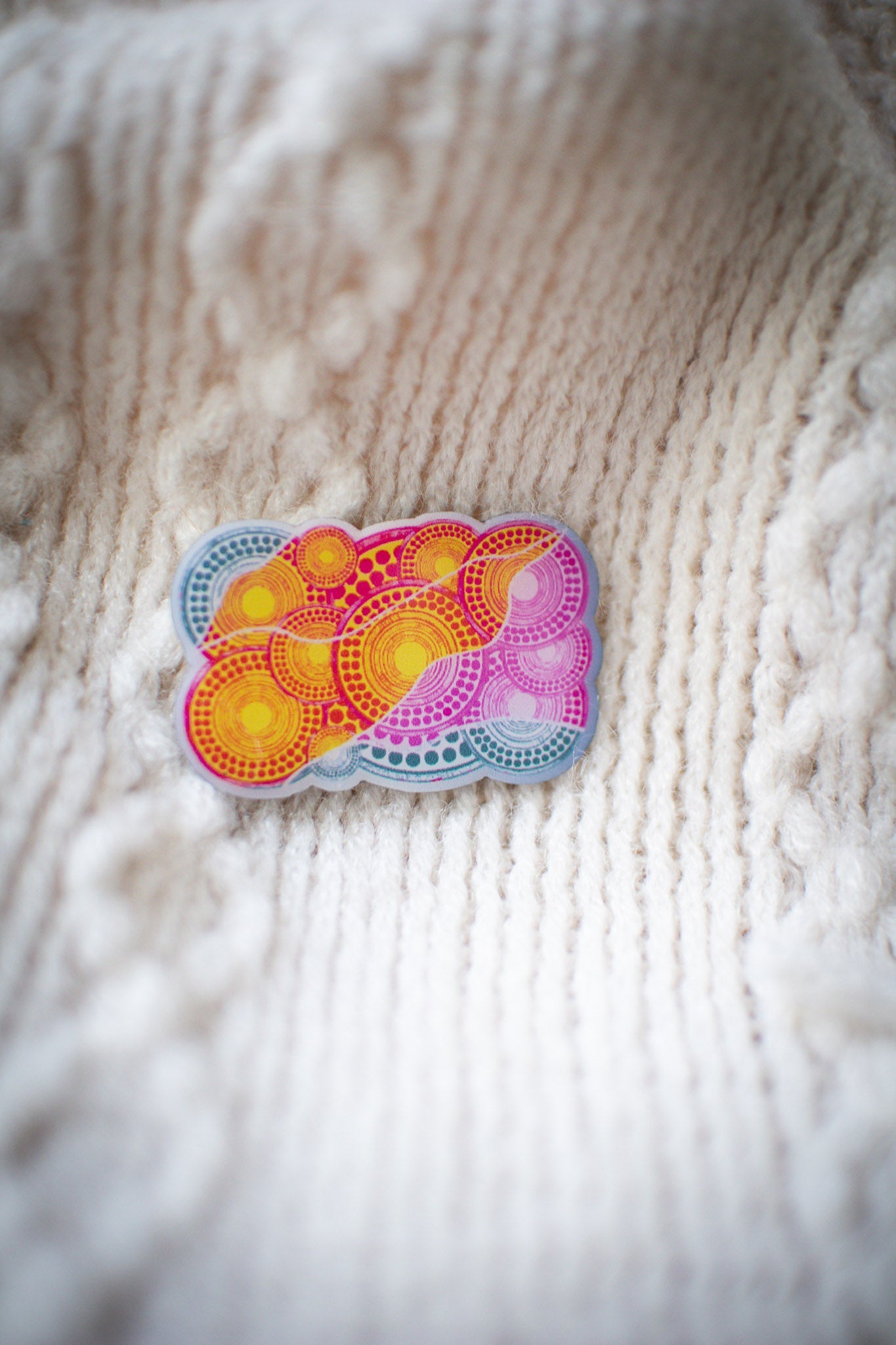 Luwaytini pin with detailed pink and yellow Indigenous artwork design, resting on a white knit sweater showing textured fabric in the background, available at the RMIT Store.