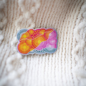 Luwaytini pin with detailed pink and yellow Indigenous artwork design, resting on a white knit sweater showing textured fabric in the background, available at the RMIT Store.