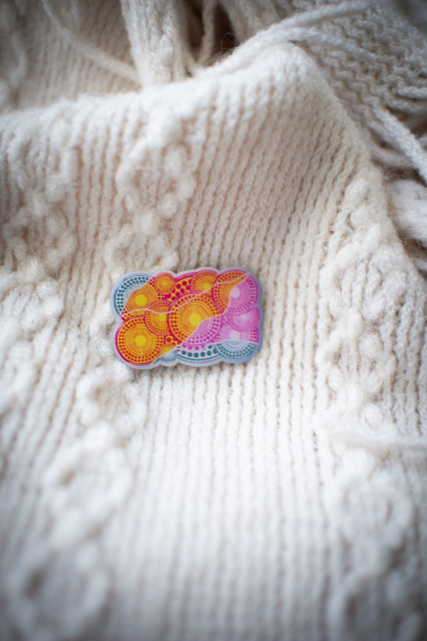 Luwaytini pin with detailed pink and yellow Indigenous artwork design, resting on a white knit sweater showing textured fabric in the background, available at the RMIT Store.