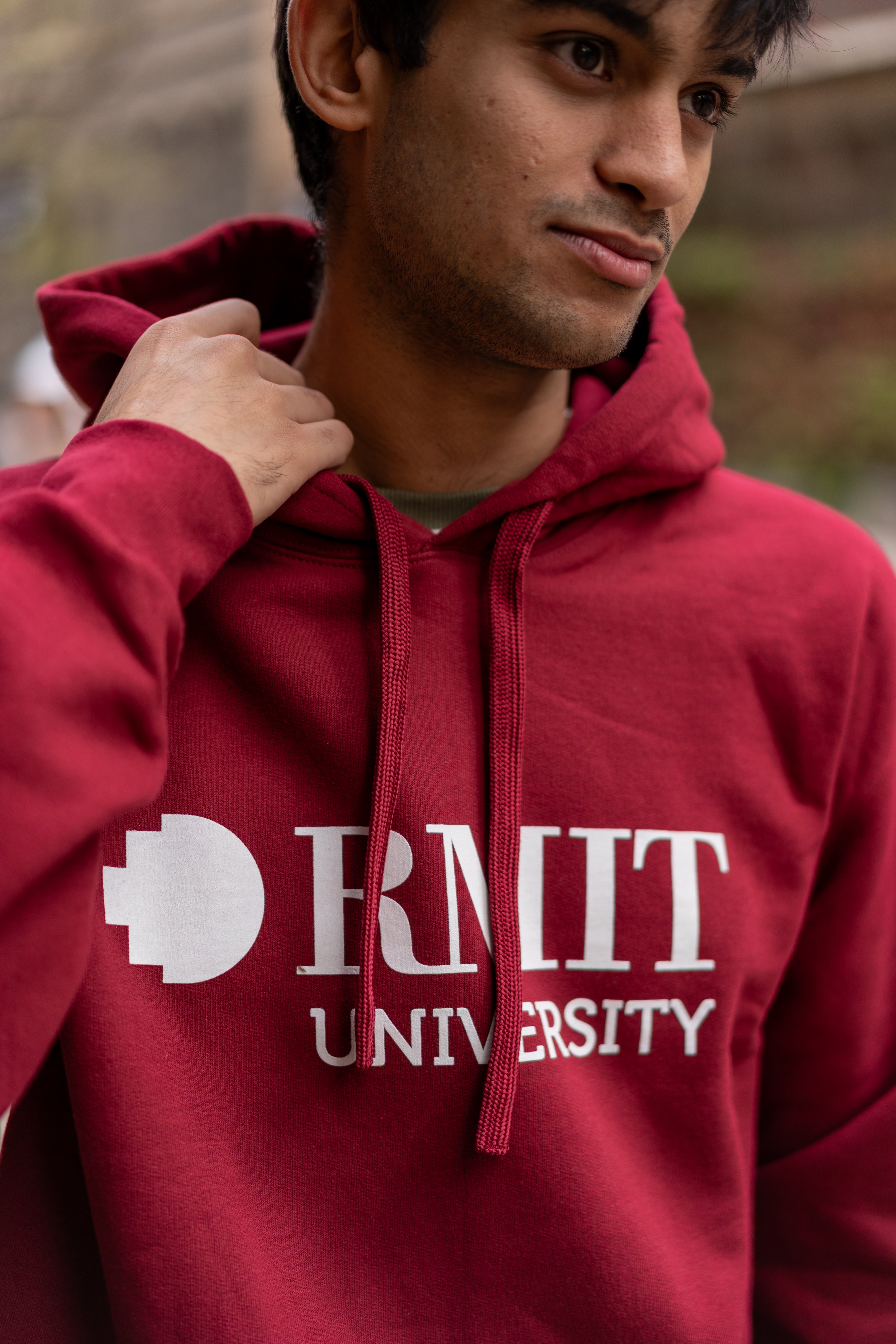 Close-up detail of a male model wearing the Lygon Hoodie in Garnet red featuring large white RMIT University logo print across the chest, matching garnet drawstrings and heavyweight fleece fabric, photographed outdoors with blurred urban background, available at the RMIT Store.