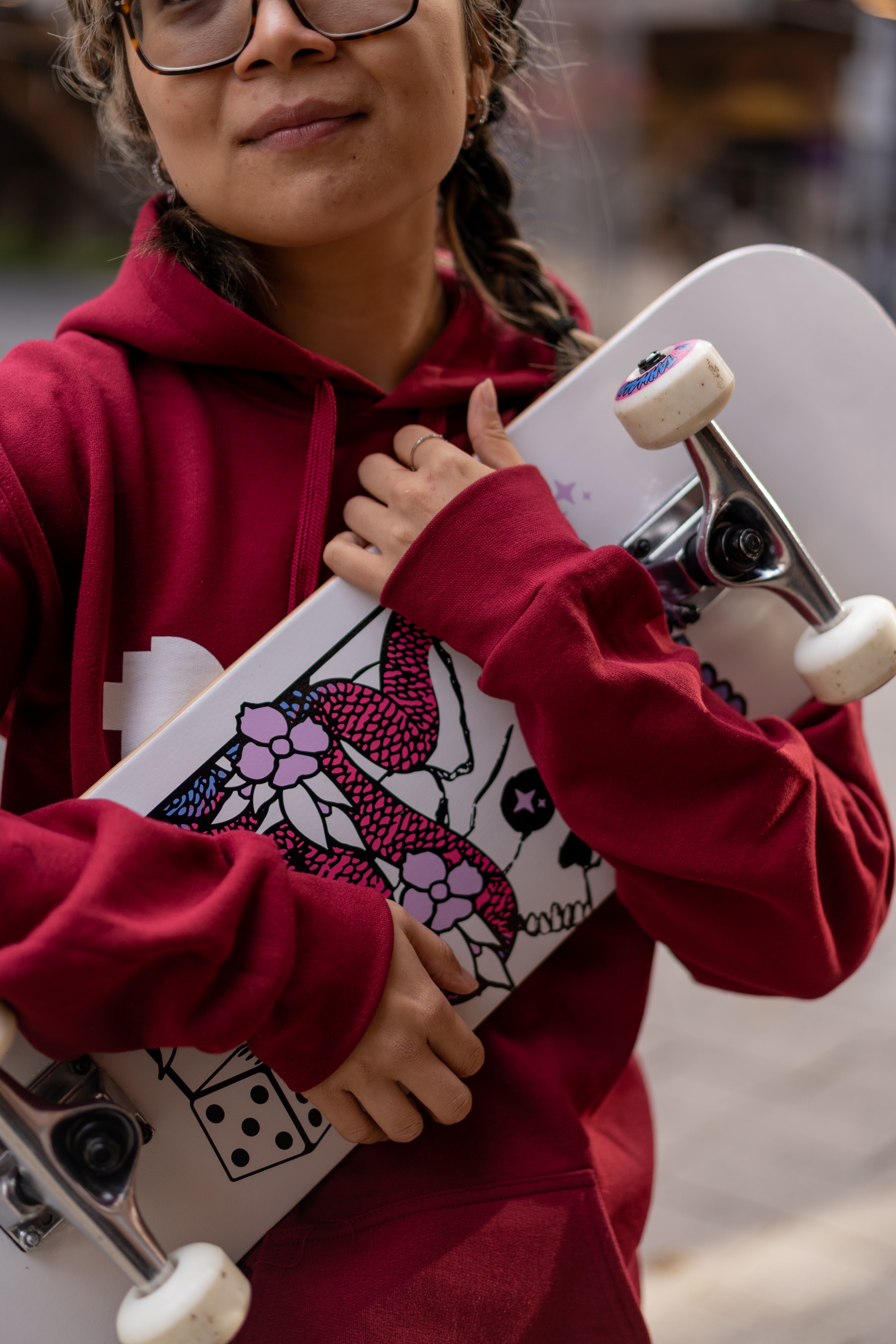 Close-up of a female model wearing the Lygon Hoodie in Garnet red with large white RMIT University logo print on the front, holding a white skateboard featuring pink floral skull artwork, black dice illustration and silver trucks with white wheels, photographed outdoors in an urban setting, available at the RMIT Store.