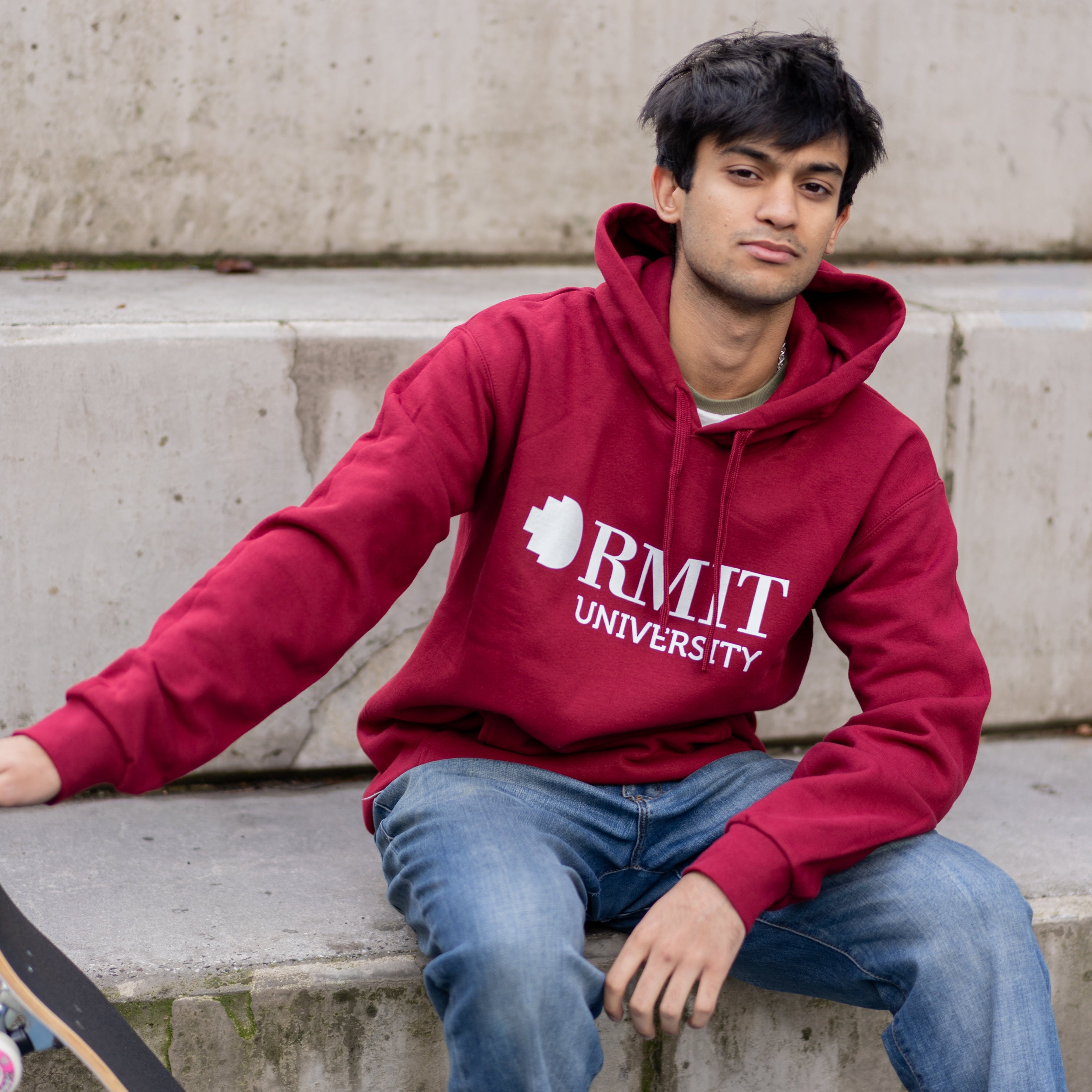 Male model wearing the Lygon Hoodie in Garnet red with white RMIT University logo print, seated on concrete steps beside a skateboard, styled with blue jeans and white sneakers in an outdoor urban setting, available at the RMIT Store.
