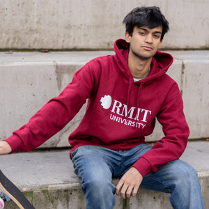 Male model wearing the Lygon Hoodie in Garnet red with white RMIT University logo print, seated on concrete steps beside a skateboard, styled with blue jeans and white sneakers in an outdoor urban setting, available at the RMIT Store.