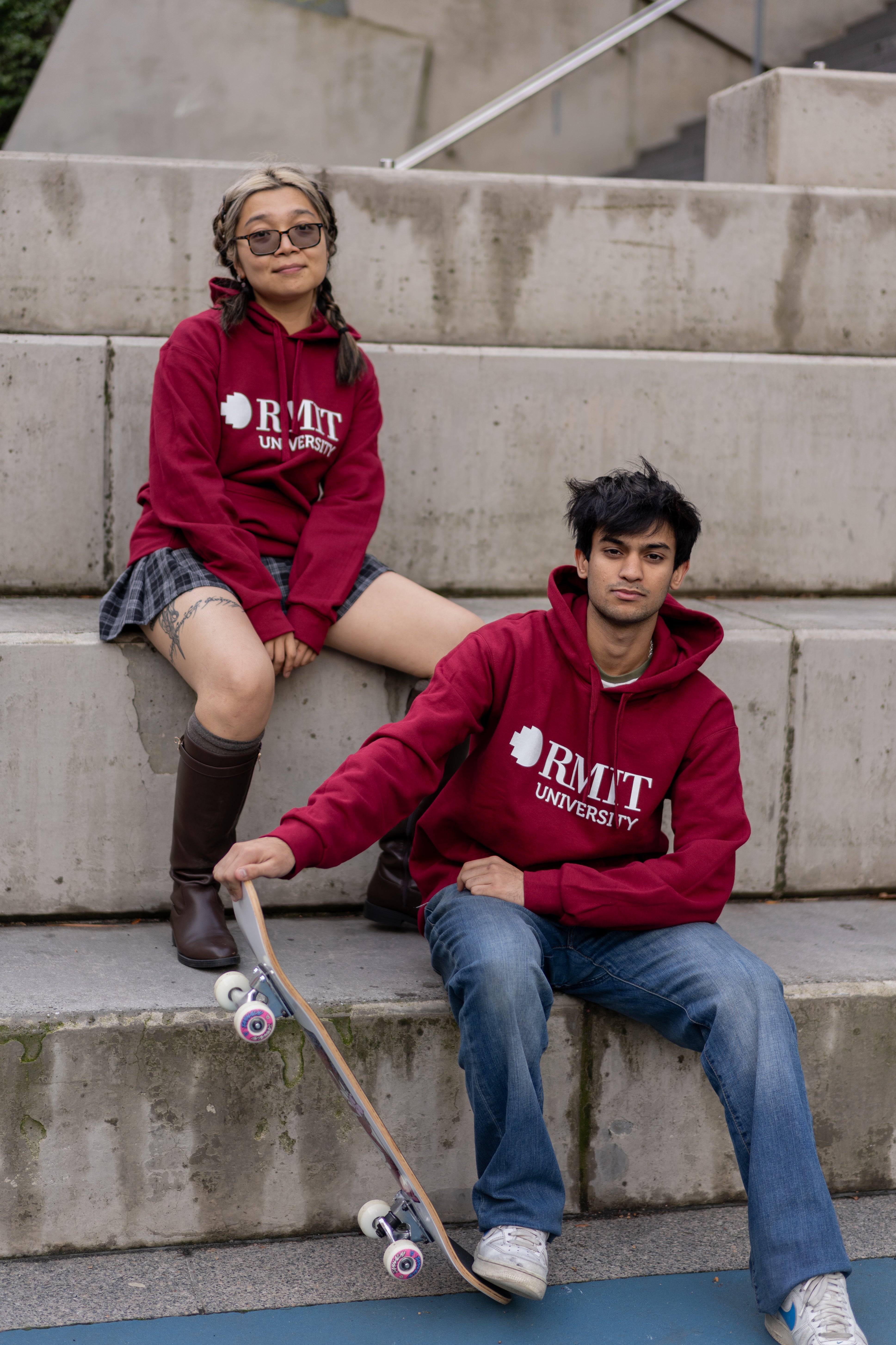 Female and male models wearing matching Lygon Hoodies in Garnet red with white RMIT University chest logos, seated on concrete steps beside a skateboard in an urban outdoor setting, styled with casual denim and streetwear-inspired outfits, available at the RMIT Store.