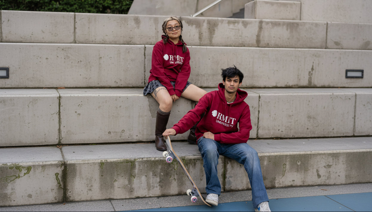 Female and male models wearing matching Lygon Hoodies in Garnet red with white RMIT University chest logos, seated on concrete steps beside a skateboard in an urban outdoor setting, styled with casual denim and streetwear-inspired outfits, available at the RMIT Store.