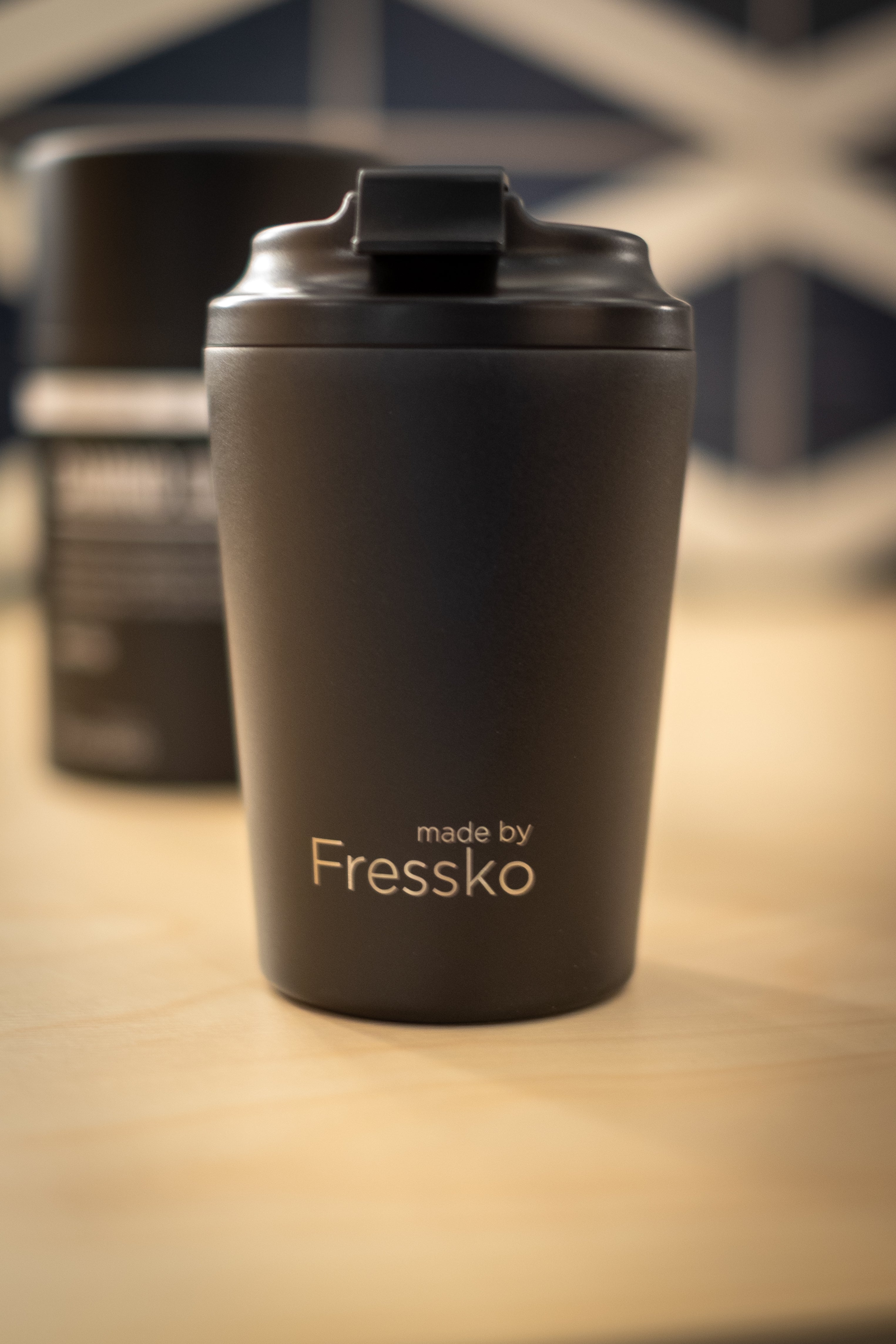 Made by Fressko x RMIT Reusable Cup in matte black with flip-lock lid, displayed upright on a wooden surface at RMIT Store, showing silver “made by Fressko” branding printed on the front, smooth powder-coated finish, rounded tapered shape, and a softly blurred second cup positioned in the background.