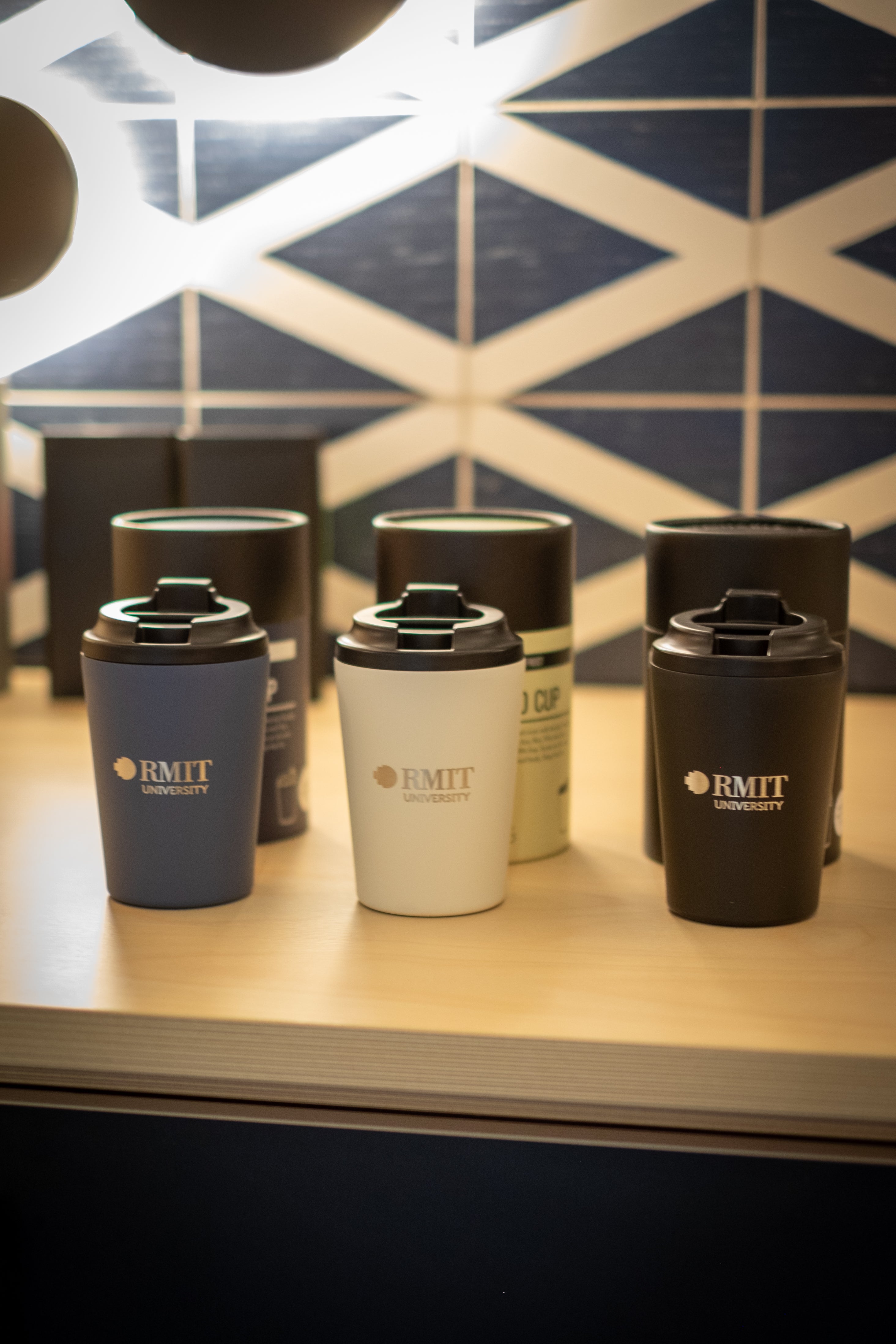 Made by Fressko x RMIT Reusable Cup range displayed on a wooden shelf at RMIT Store, showing three upright cups in navy blue, white, and black with printed RMIT University logos on the front, black flip-lock lids, and additional cups positioned behind against a geometric tiled wall.