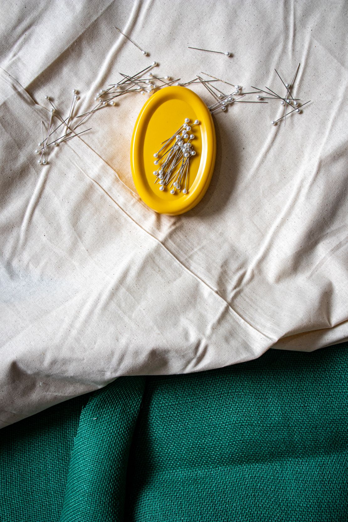 Top view of the yellow magnetic pin holder with multiple pearl-headed pins scattered loosely across the surface, displayed on a beige fabric laid over a piece of teal fabric, available from the RMIT Store.