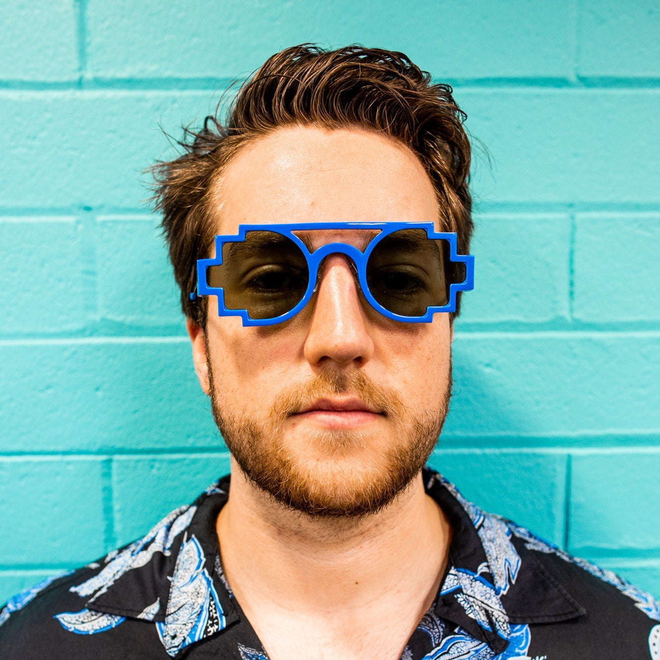 Male model wearing a short-sleeved floral shirt, paired with sky blue pixelated sunglasses. The model is positioned facing forward against a solid blue studio background, with a relaxed expression and natural lighting for the RMIT Store.