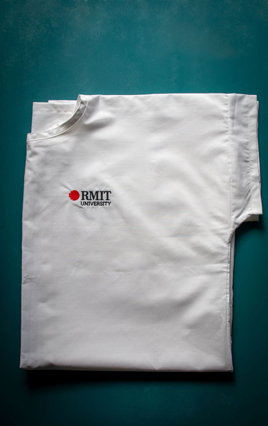 White Medical Lab Gown from the RMIT Store, folded neatly into a near-square shape. The gown features short sleeves, a round collar, and the RMIT University logo in black and red near the collar. The gown is made from lightweight, smooth fabric. The background is a neutral, light surface, highlighting the compact fold and clean, professional design of the gown.