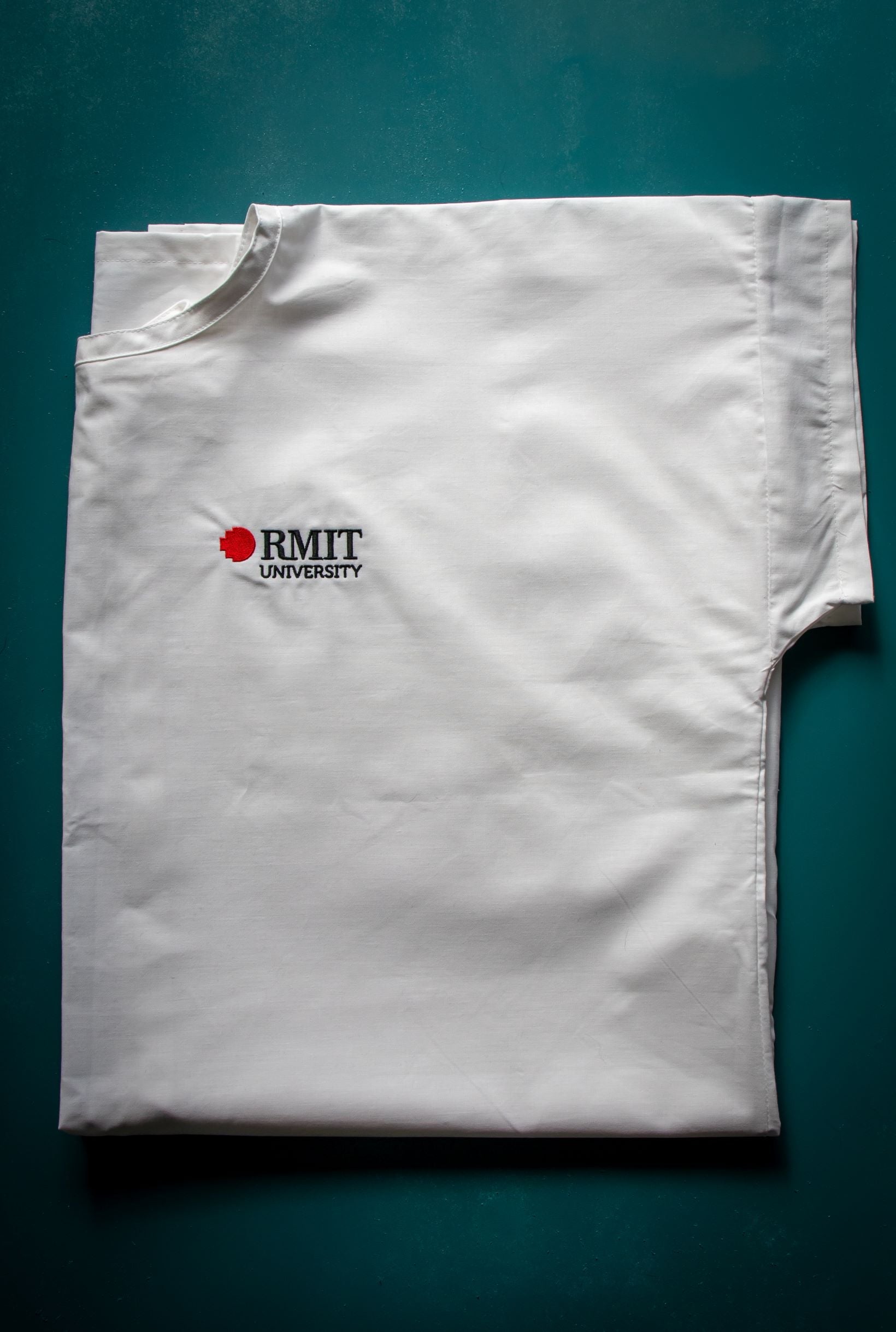 Folded white Medical Lab Gown from the RMIT Store displayed on a plain surface, showing the front side with one visible short sleeve and a round collar. The RMIT University logo, printed in black and red, appears clearly on the chest area.
