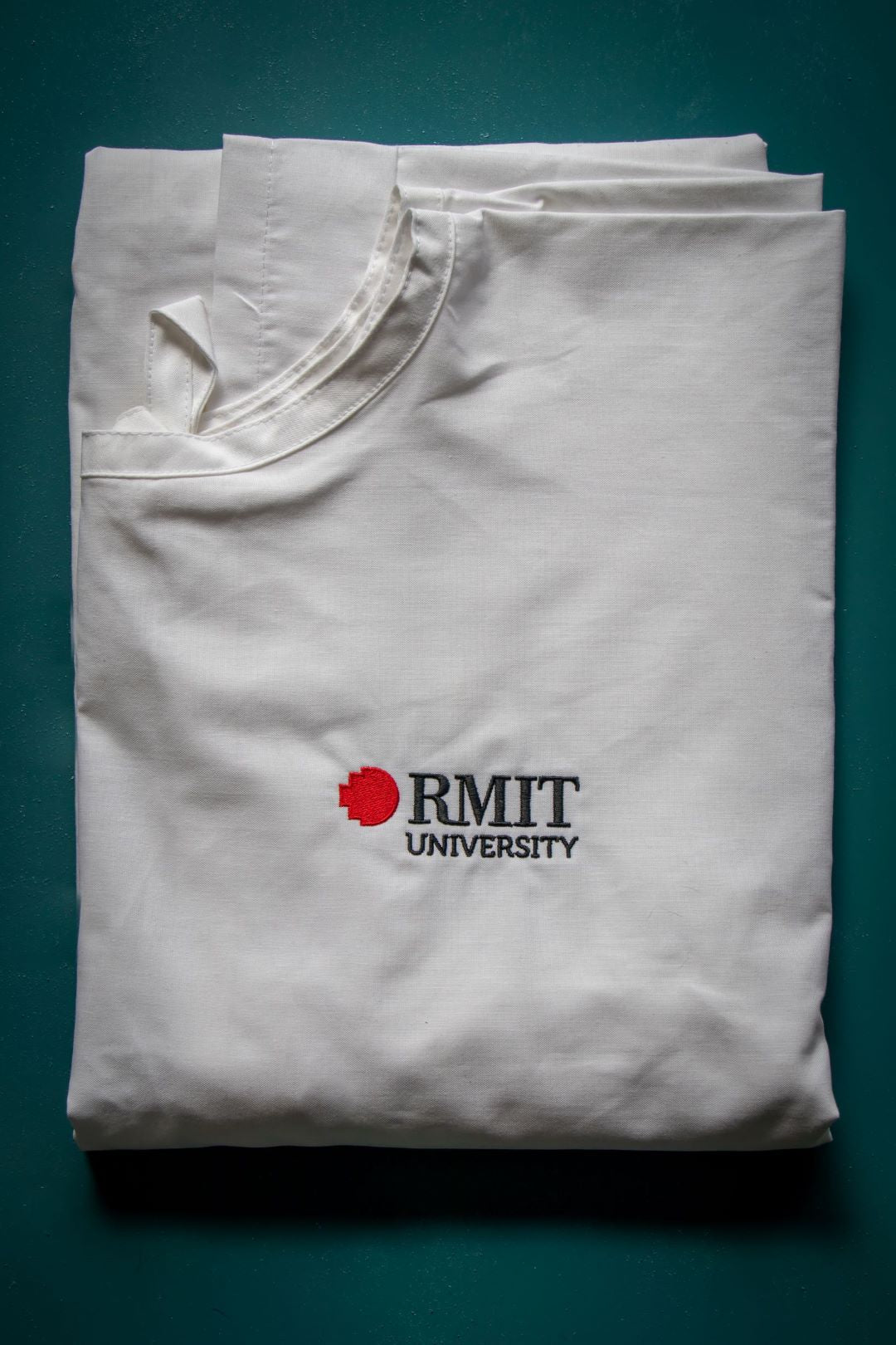 White RMIT University medical lab gown with embroidered logo from the RMIT Store, showcased folded on a teal background, illustrating its professional and clean design.