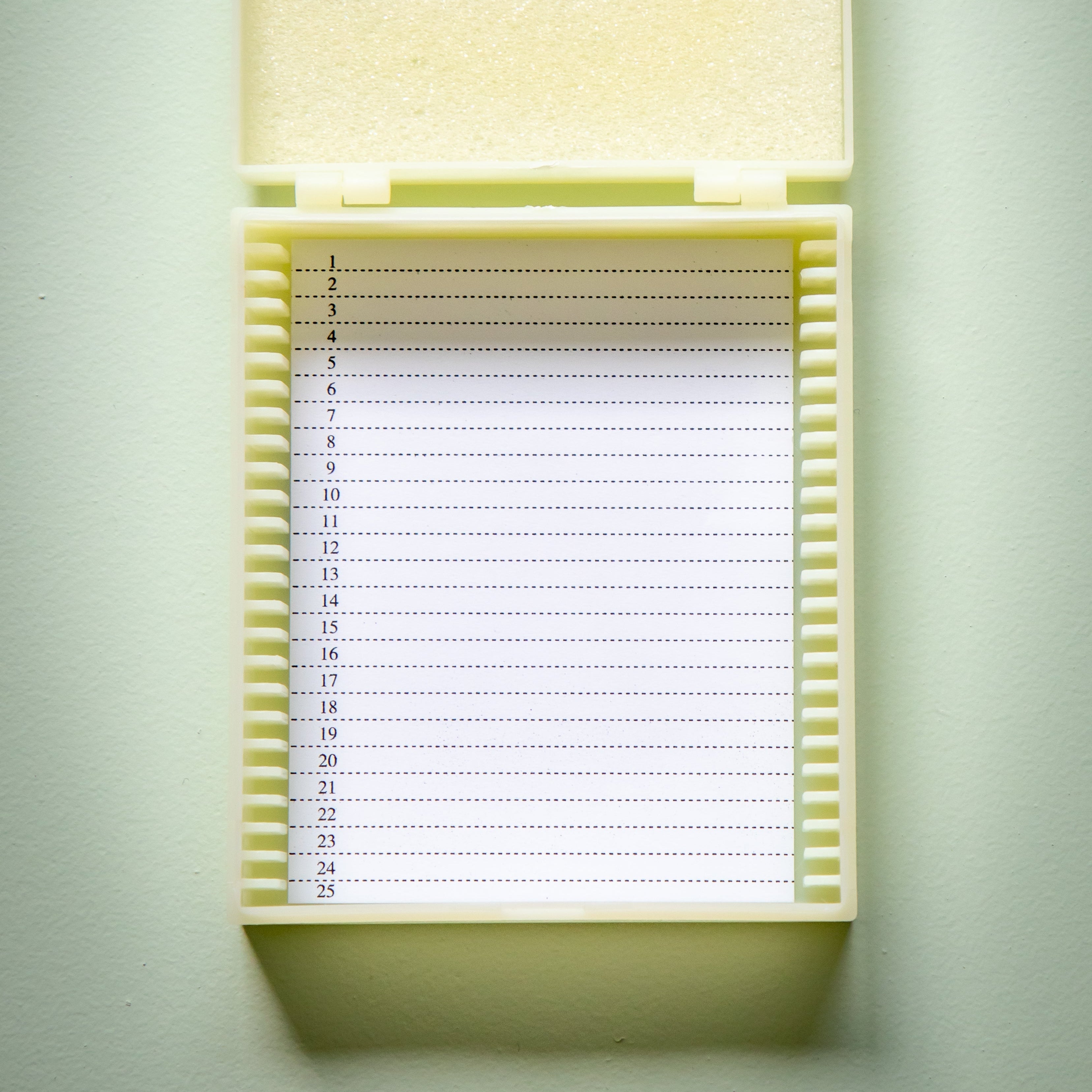 Microslide Plastic Case by Livingstone shown open with the hinged lid raised, revealing numbered storage slots for 25 microscope slides, an internal foam insert in the lid for slide protection, and a printed numbered index from 1 to 25. The reusable off-white plastic case is photographed against a plain light background for the RMIT Store product catalogue.