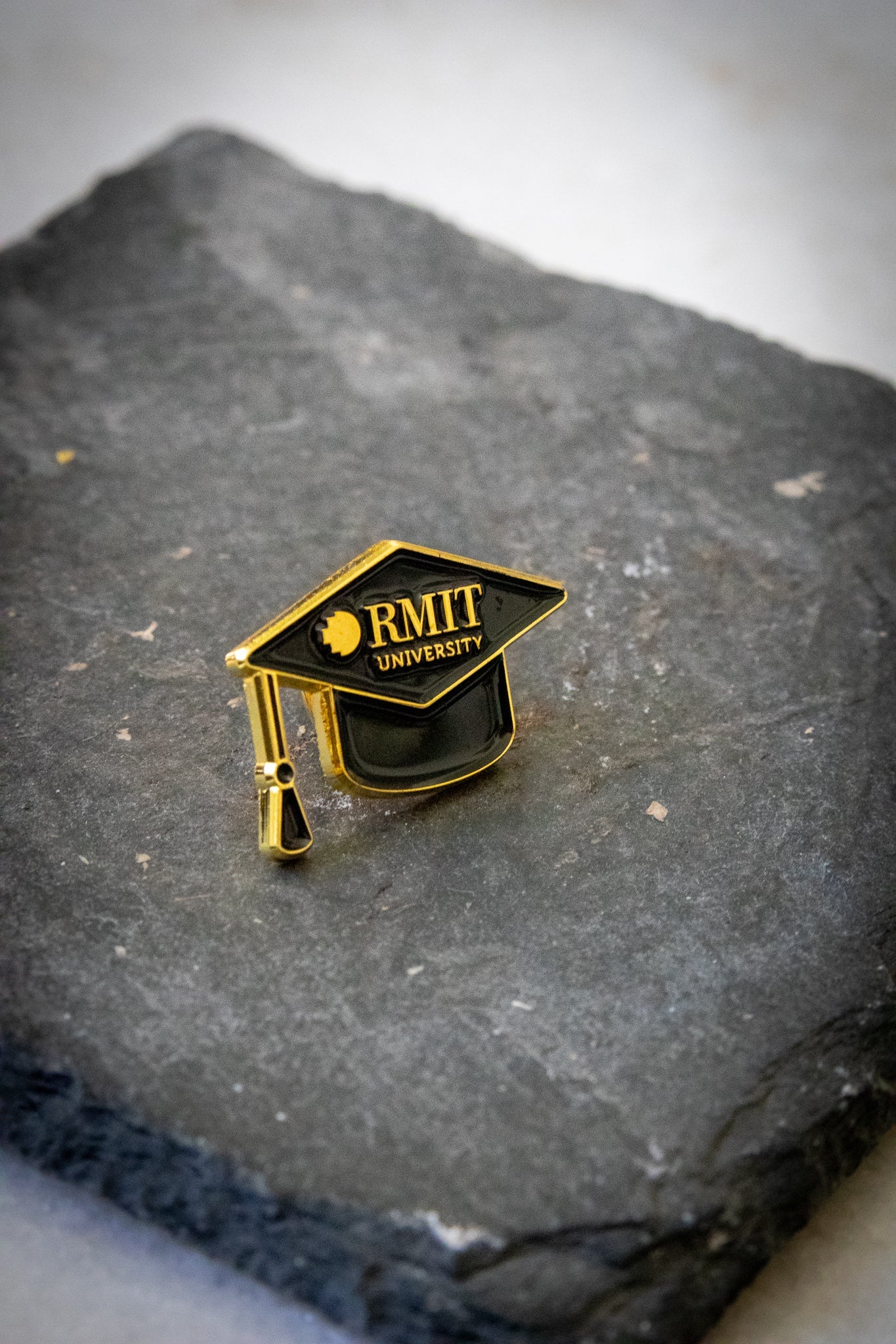 Angled front view of the mortarboard pin featuring black enamel with gold detailing, including gold RMIT branding across the front, photographed on a dark grey stone background; available at the RMIT Store.
