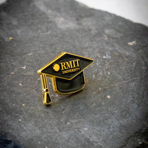 Angled front view of the mortarboard pin featuring black enamel with gold detailing, including gold RMIT branding across the front, photographed on a dark grey stone background; available at the RMIT Store.