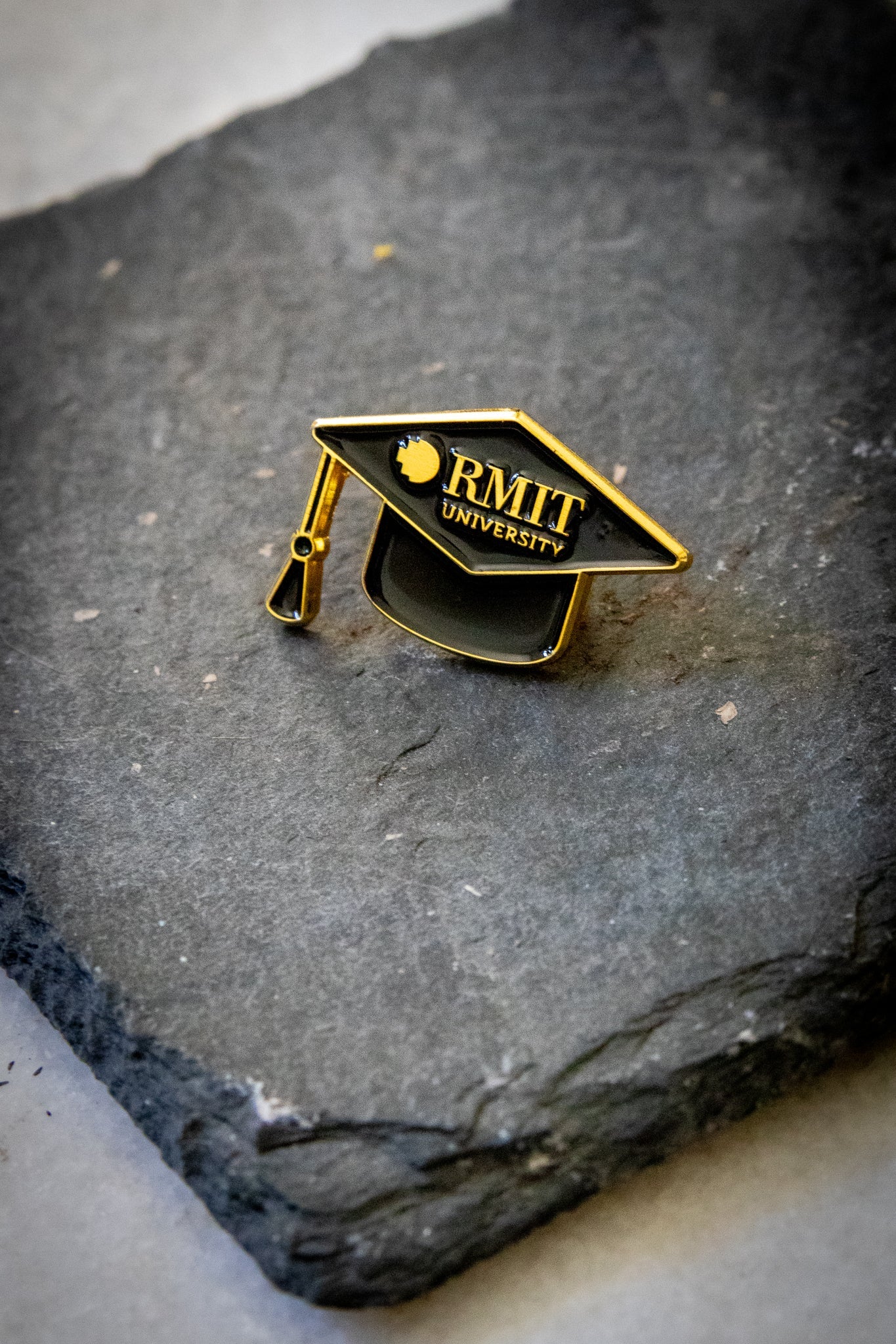 Angled front view of the mortarboard pin featuring black enamel with gold detailing, including gold RMIT branding across the front, photographed on a dark grey stone background; available at the RMIT Store.