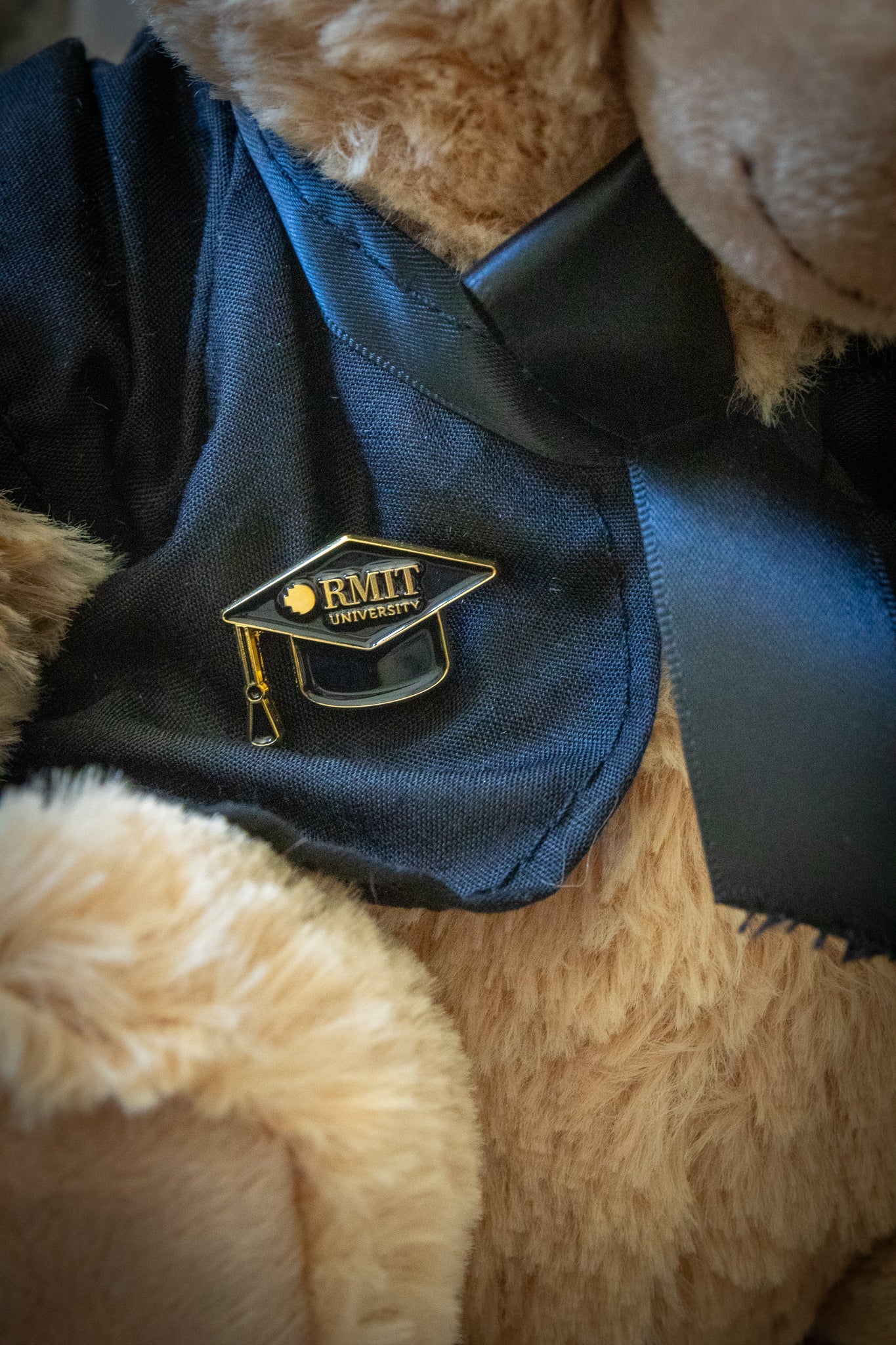 Zoomed-in view of the mortarboard pin with black enamel and gold RMIT branding, resting on the plain black gown of an RMIT Graduation Bear, highlighting the fine detailing of the pin against the soft fabric. Available at the RMIT Store.