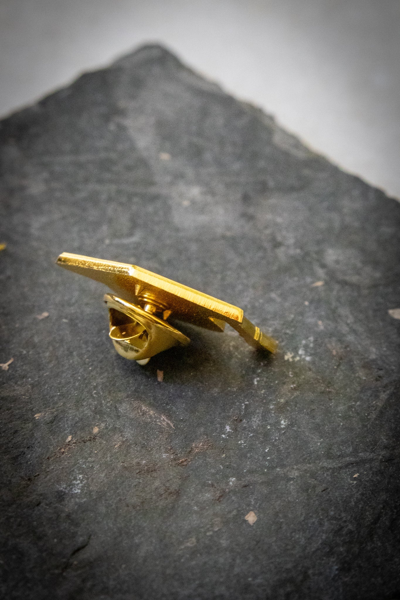 Side view of a mortarboard pin with gold enamel on the back and butterfly clasp, resting on a dark grey stone ground, showing detailed pin mechanism; product available at the RMIT Store.