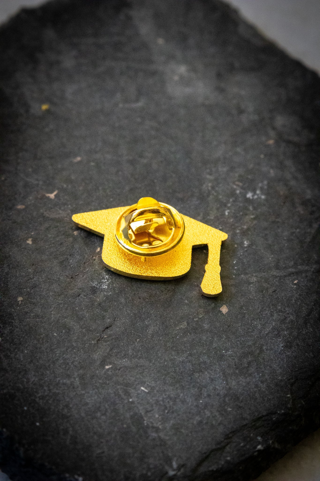 Rear view of the mortarboard pin showing smooth gold enamel surface and butterfly clasp fastener, photographed on a dark grey stone background; product available at the RMIT Store