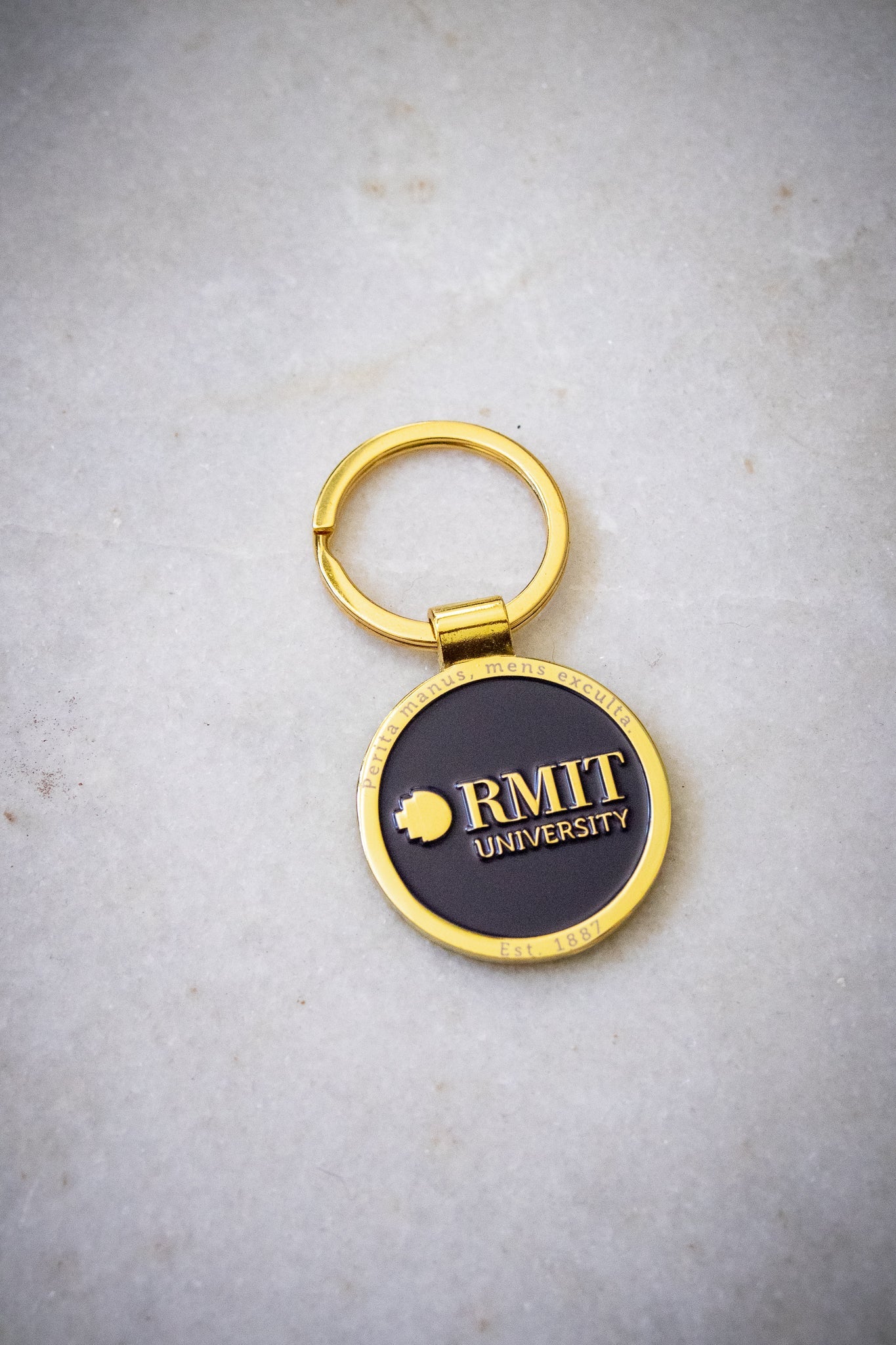 Angled front view of the RMIT motto lapel keyring, featuring a navy centre with a gold outer edge and gold RMIT branding, attached to a gold circular handle, displayed on a light background, available at the RMIT Store.