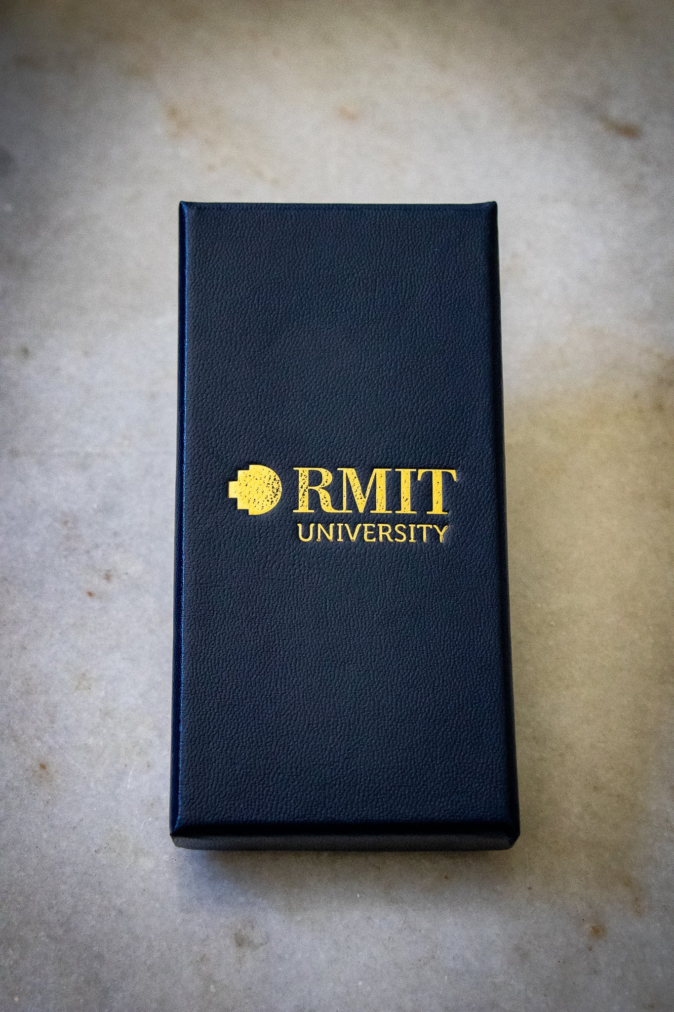 Rectangular navy gift box for the RMIT motto lapel keyring, featuring gold RMIT branding on the lid, laid flat on a light background, available at the RMIT Store.