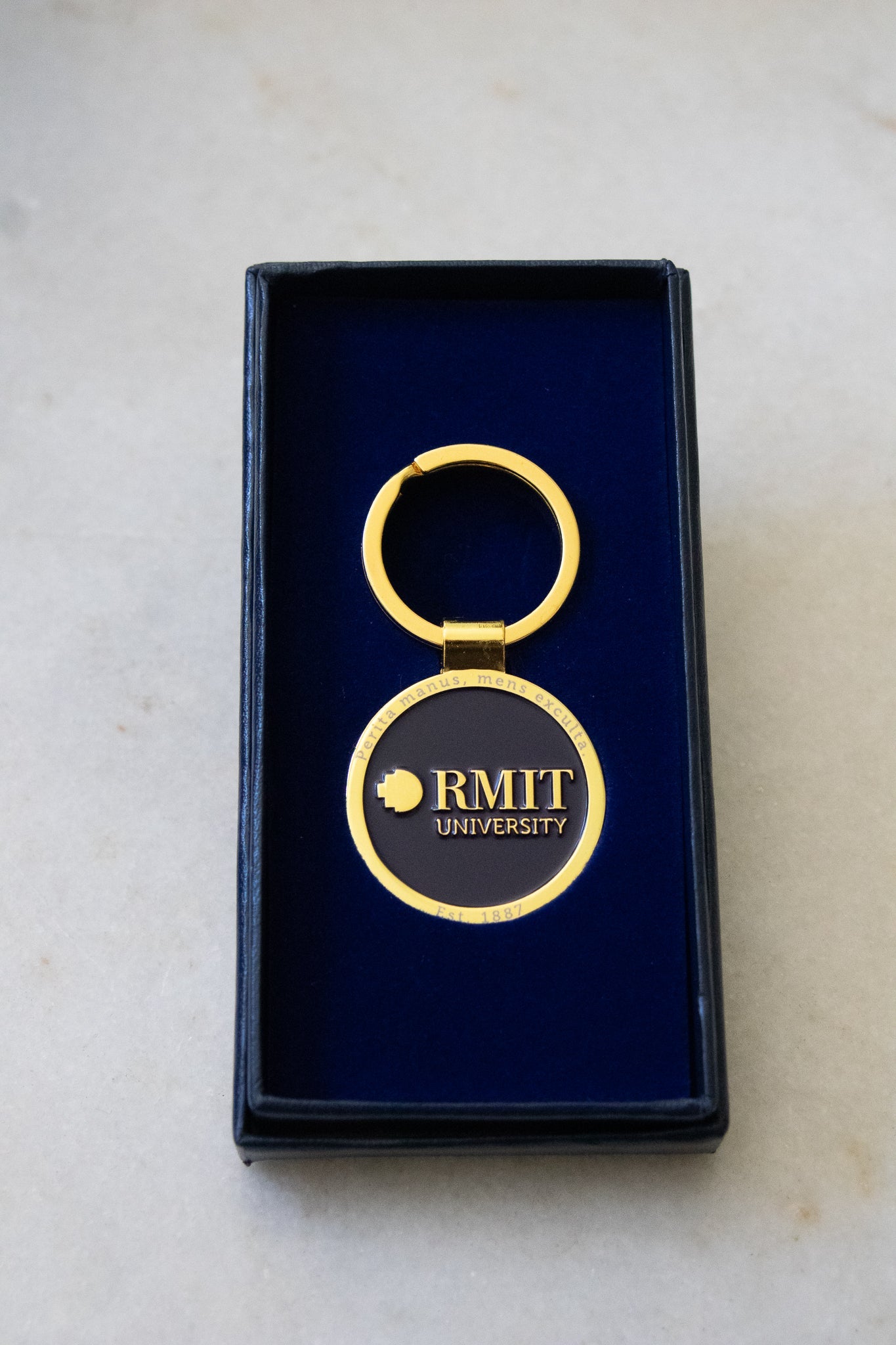 Rectangular navy gift box with the lid placed underneath, containing the RMIT motto lapel keyring inside. The keyring features a navy centre with a gold outer edge and a gold circular handle, set against a light background, available at the RMIT Store.