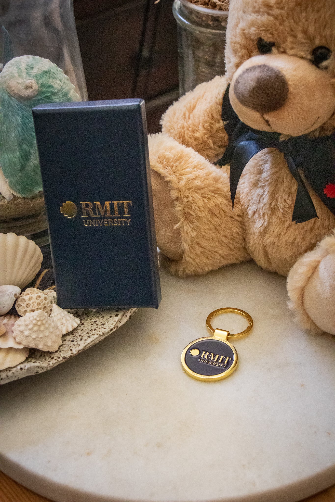 RMIT motto lapel keyring and gift box displayed in front of a graduation bear. The rectangular navy box with gold RMIT branding sits on the left, while the navy keyring with a gold outer edge and gold circular handle is on the right, available at the RMIT Store.