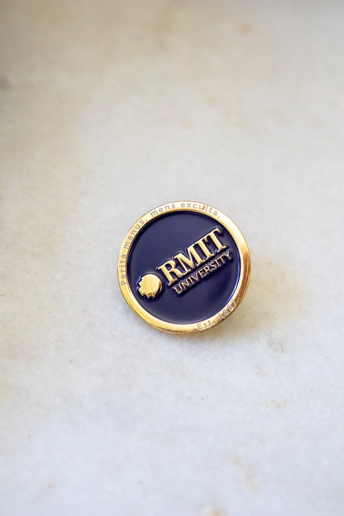 Close-up view of the RMIT motto lapel pin featuring a gold outer edge inscribed with the RMIT motto, a navy centre, and gold RMIT branding in the middle, available at the RMIT Store.