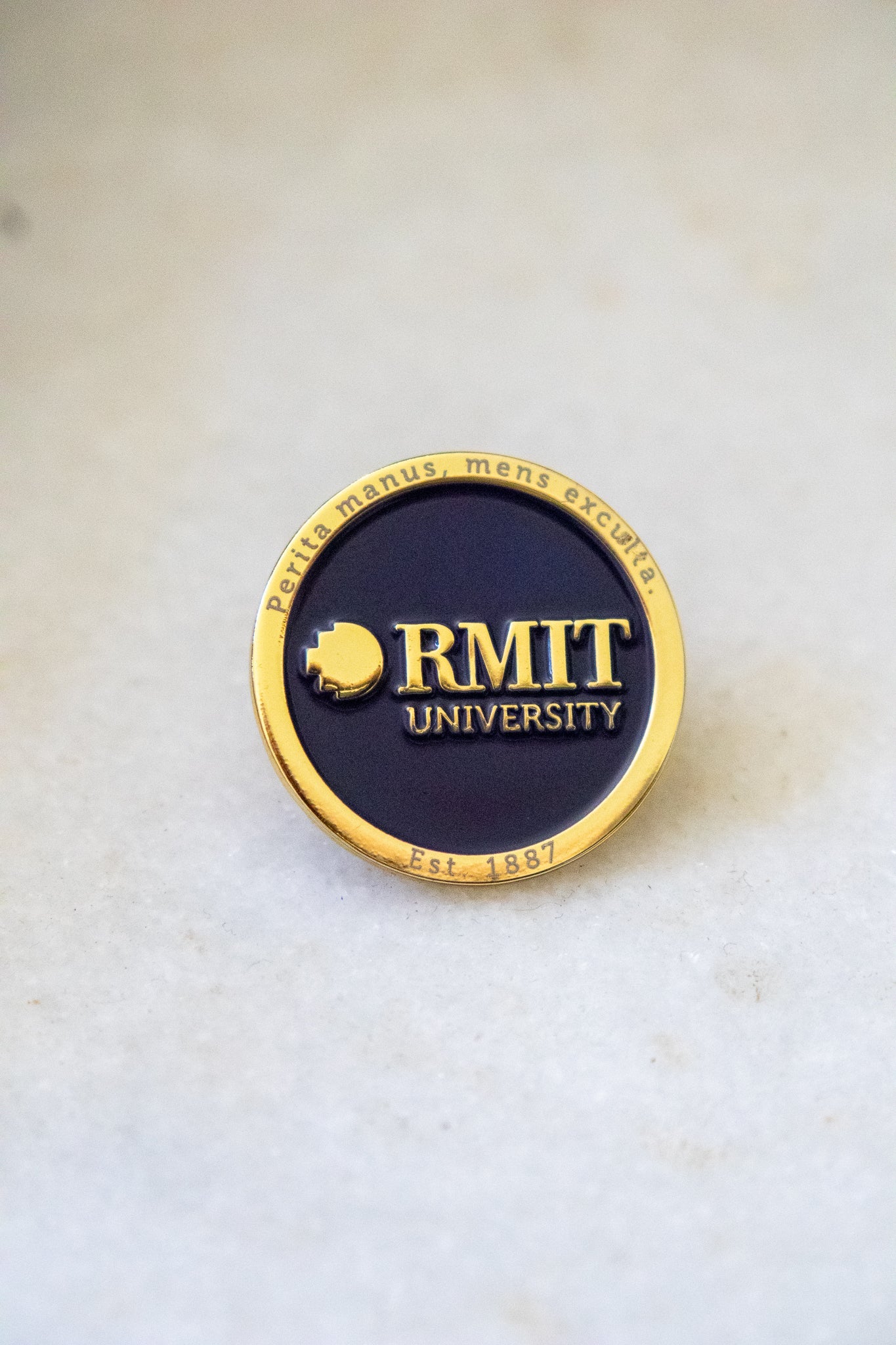 Close-up view of the RMIT motto lapel pin featuring a gold outer edge inscribed with the RMIT motto, a navy centre, and gold RMIT branding in the middle, available at the RMIT Store.