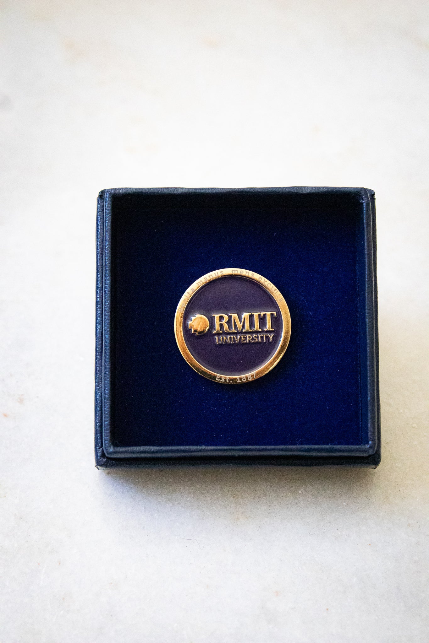 RMIT motto lapel pin displayed inside a square navy gift box, with the box lid placed beneath it. The pin features a navy centre with a gold outer edge and gold RMIT branding in the middle, available at the RMIT Store.