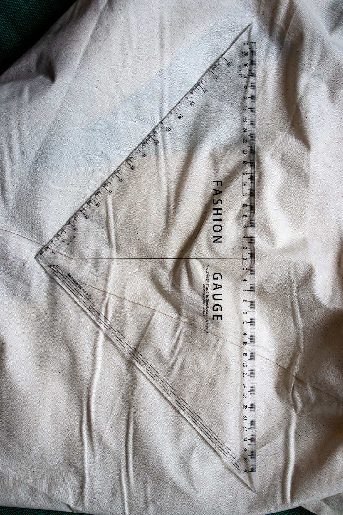 Top-down vertical view of the Multi Scale Ruler DM4 from the RMIT Store, displaying full length of clear plastic ruler with black measurement scales, lying flat on beige fabric.