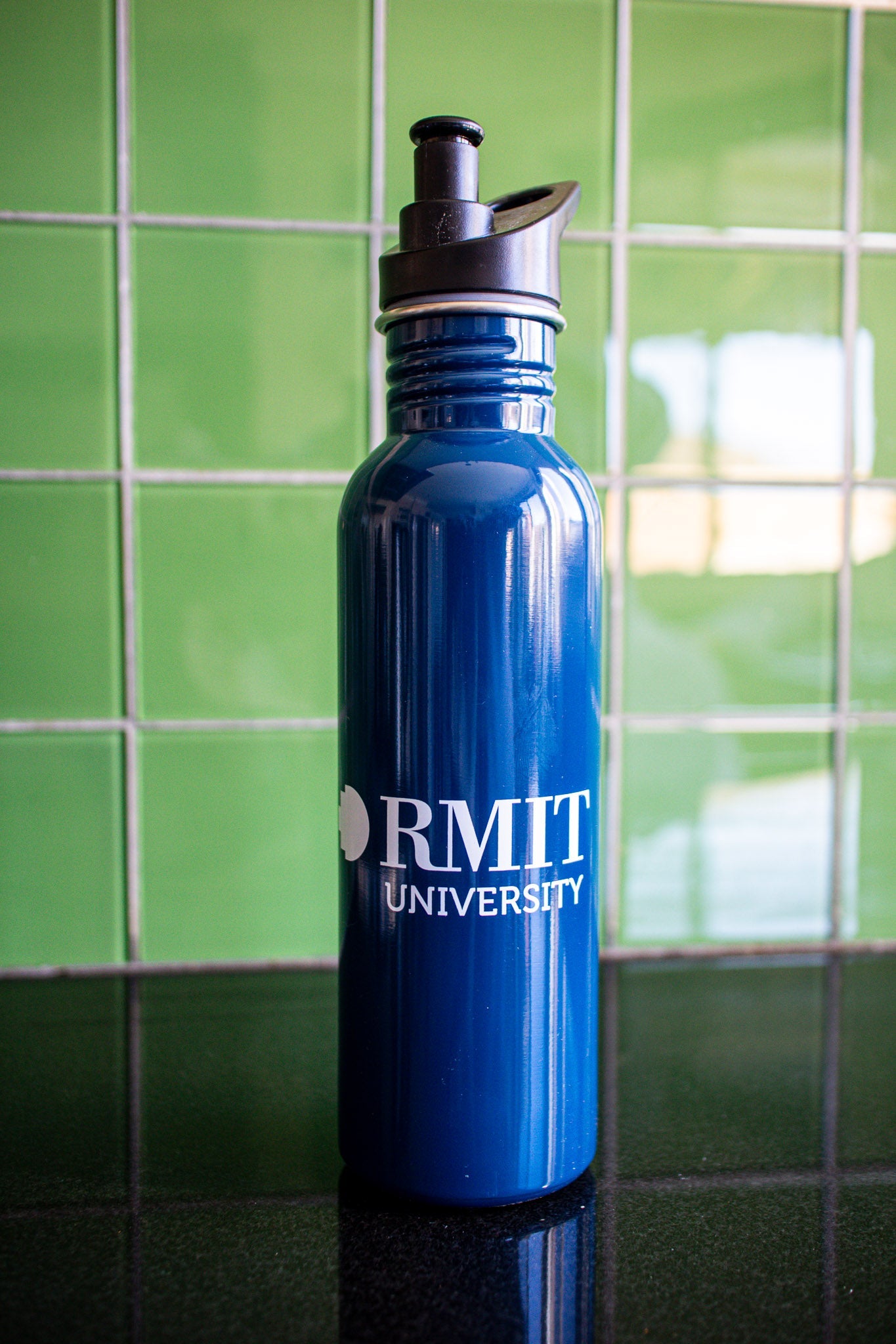Full view of the navy RMIT bottle with a white RMIT University logo centered on the body, black cap securely on the bottle, placed on a black tabletop with a green background. Available at the RMIT Store.