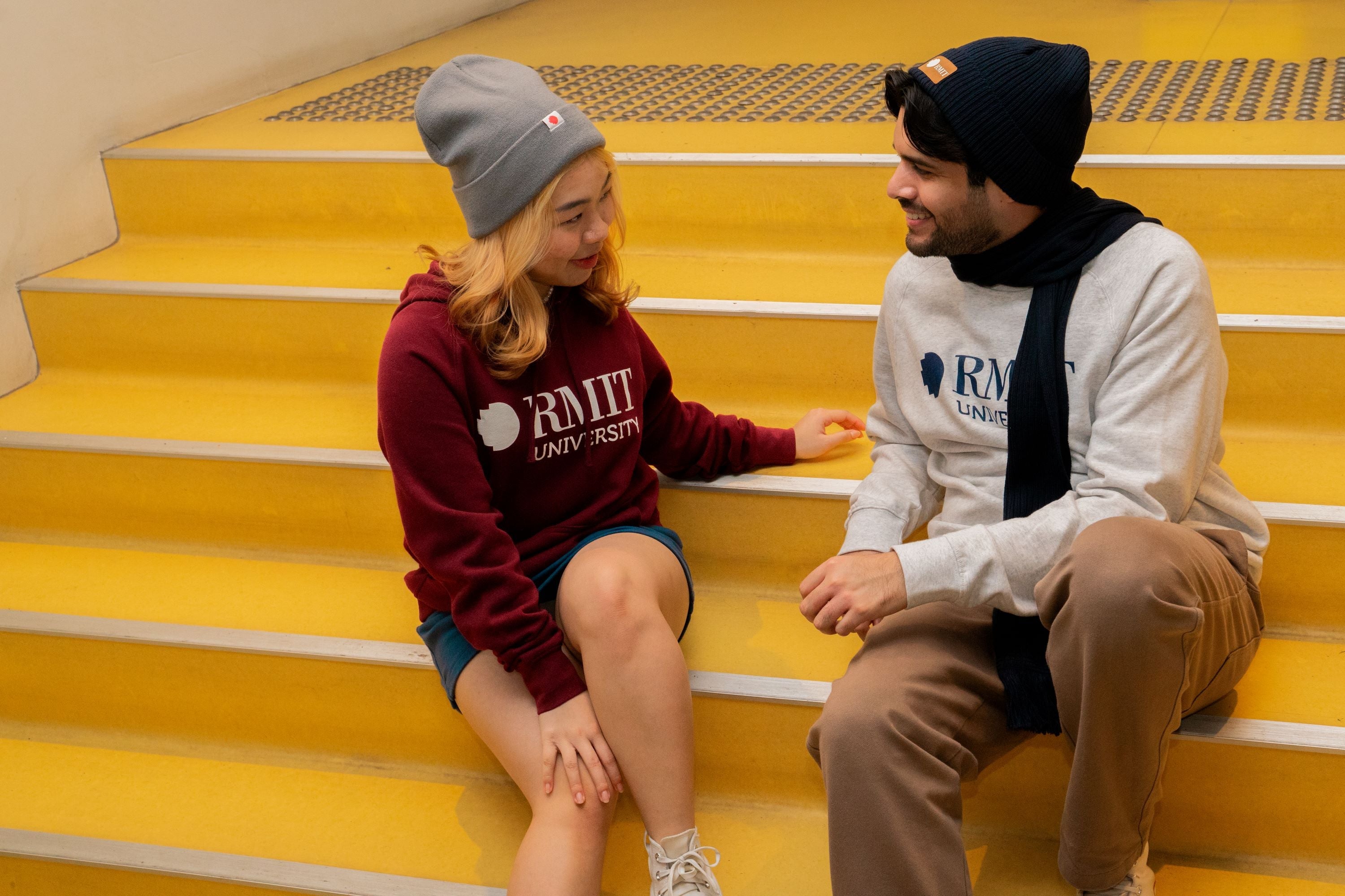 Female model seated on the left wearing the RMIT Luxe Beanie in grey paired with a red hoodie, and male model seated on the right wearing a navy knitted RMIT beanie and matching scarf paired with a white crew neck sweater. Both models are sitting on outdoor stairs in an urban setting, looking at each other with relaxed expressions. The female’s beanie features a folded cuff with a woven RMIT logo tag, while the male’s knitted beanie and scarf have a textured pattern. Available at the RMIT Store.