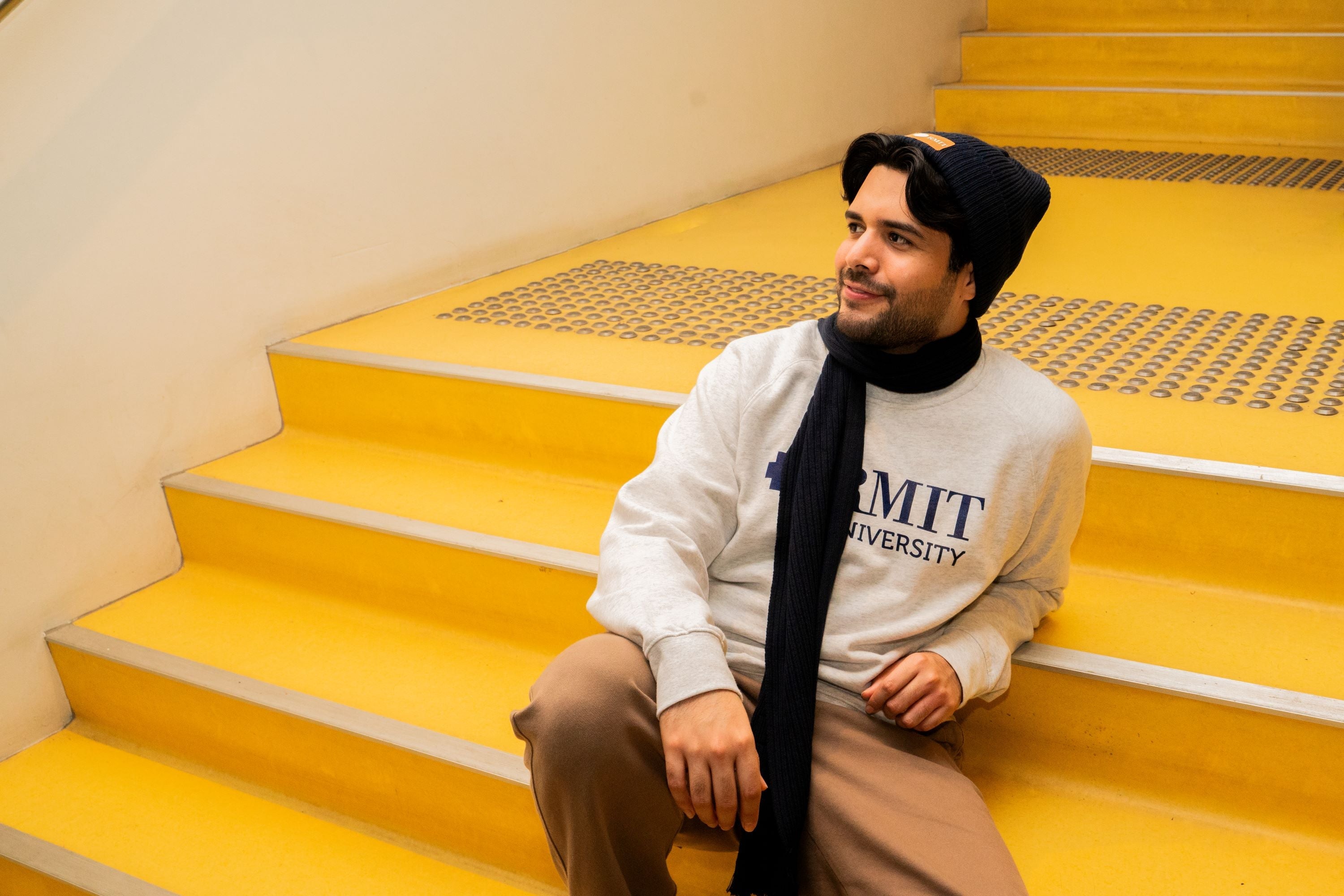 Male model seated on outdoor stairs in an urban setting, wearing the RMIT knitted beanie in navy with a brown PU patch embossed with the RMIT logo at the front. He is also wearing a matching navy knitted scarf and a white crew neck sweater. The beanie and scarf feature a textured knit pattern, styled casually for cool weather. Available at the RMIT Store.