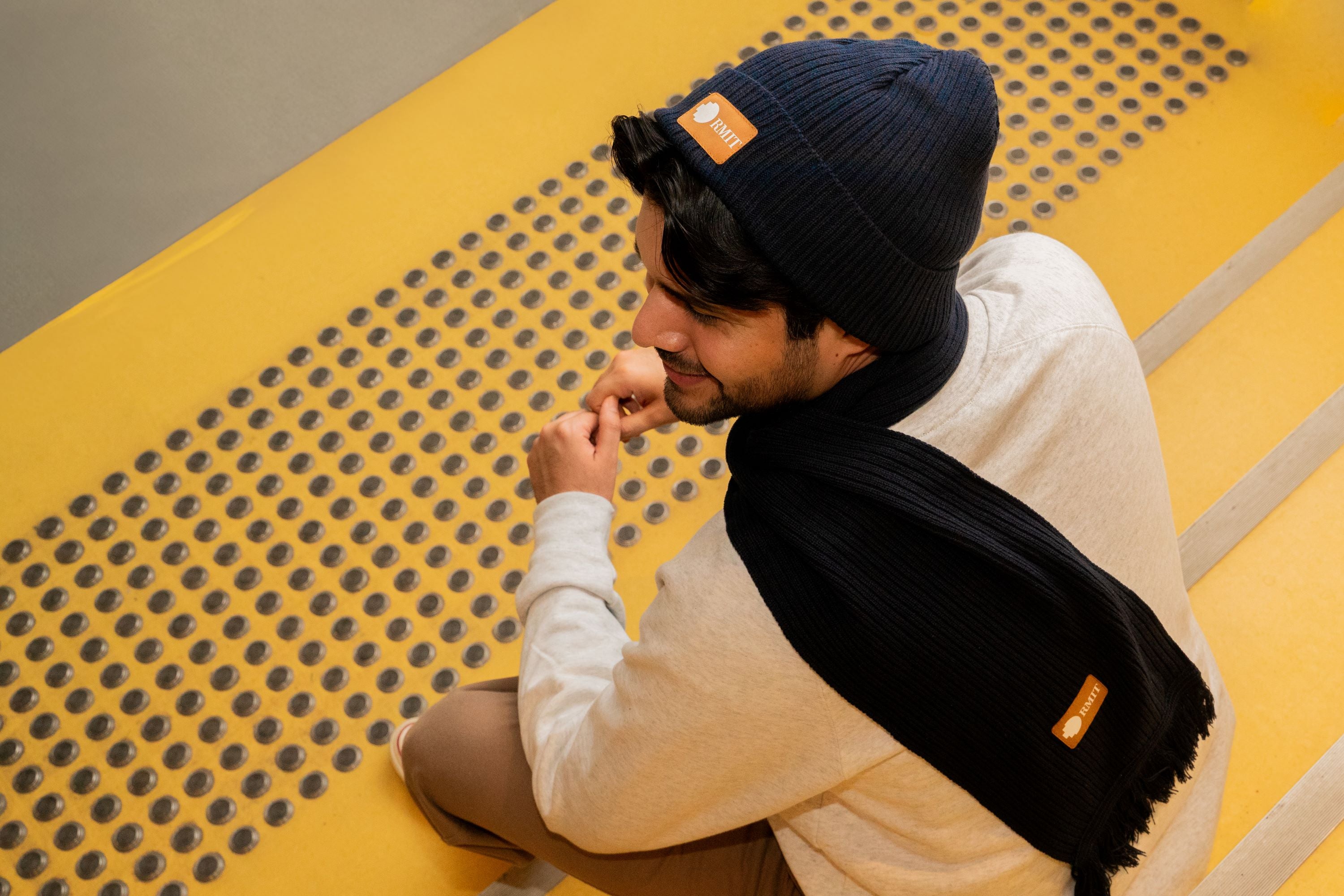 Male model looking back towards the camera while wearing the navy RMIT knitted beanie with a brown PU patch embossed with the RMIT logo, and the matching navy knitted scarf. The model is seated on outdoor stairs in an urban setting, dressed casually in a white crew neck sweater. The knit texture and details of both the beanie and scarf are clearly visible. Available at the RMIT Store.